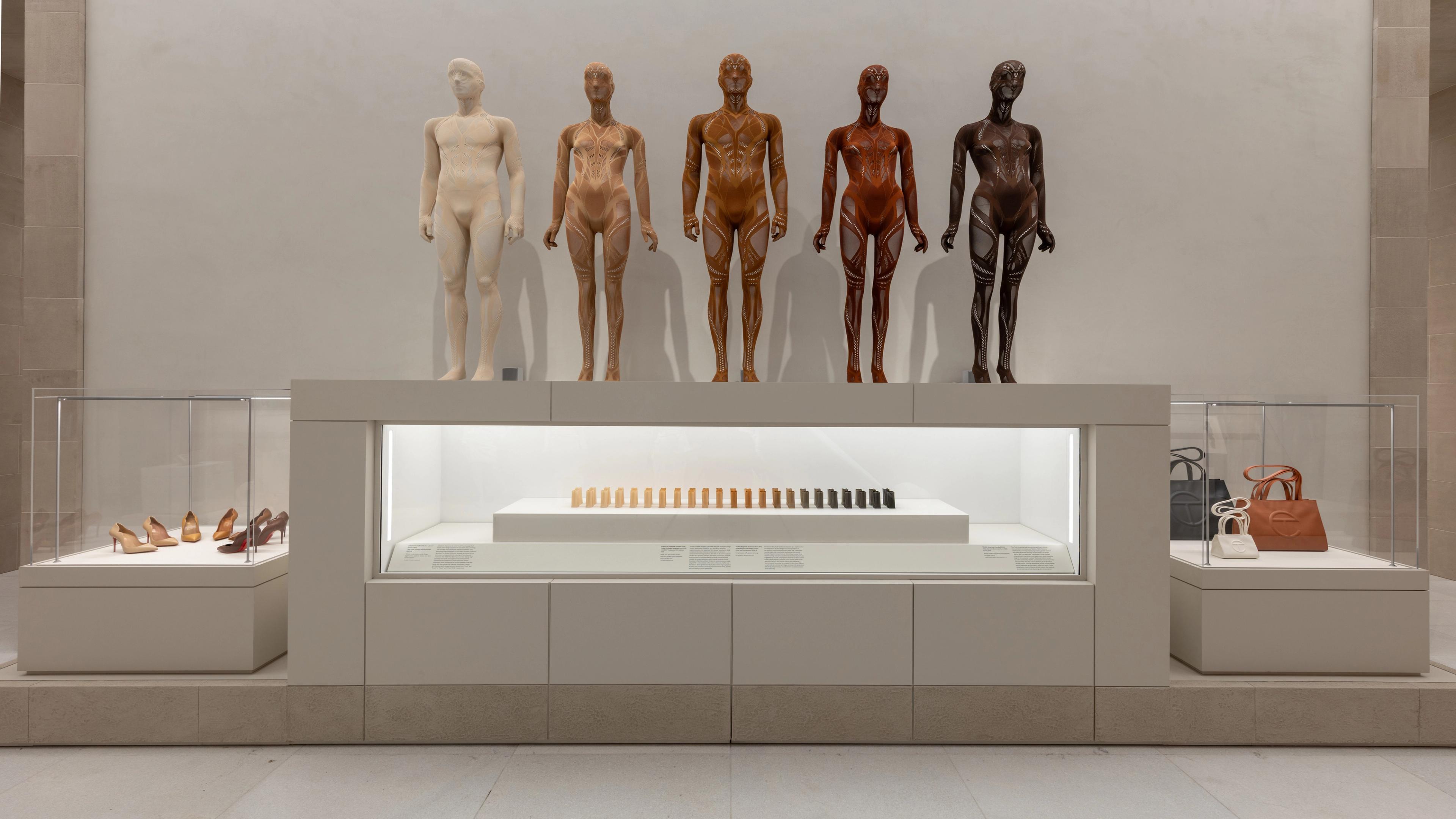 Five diverse mannequins in skin tones stand above a display of matching cosmetics. Shoes and handbags are showcased in glass cases on each side.