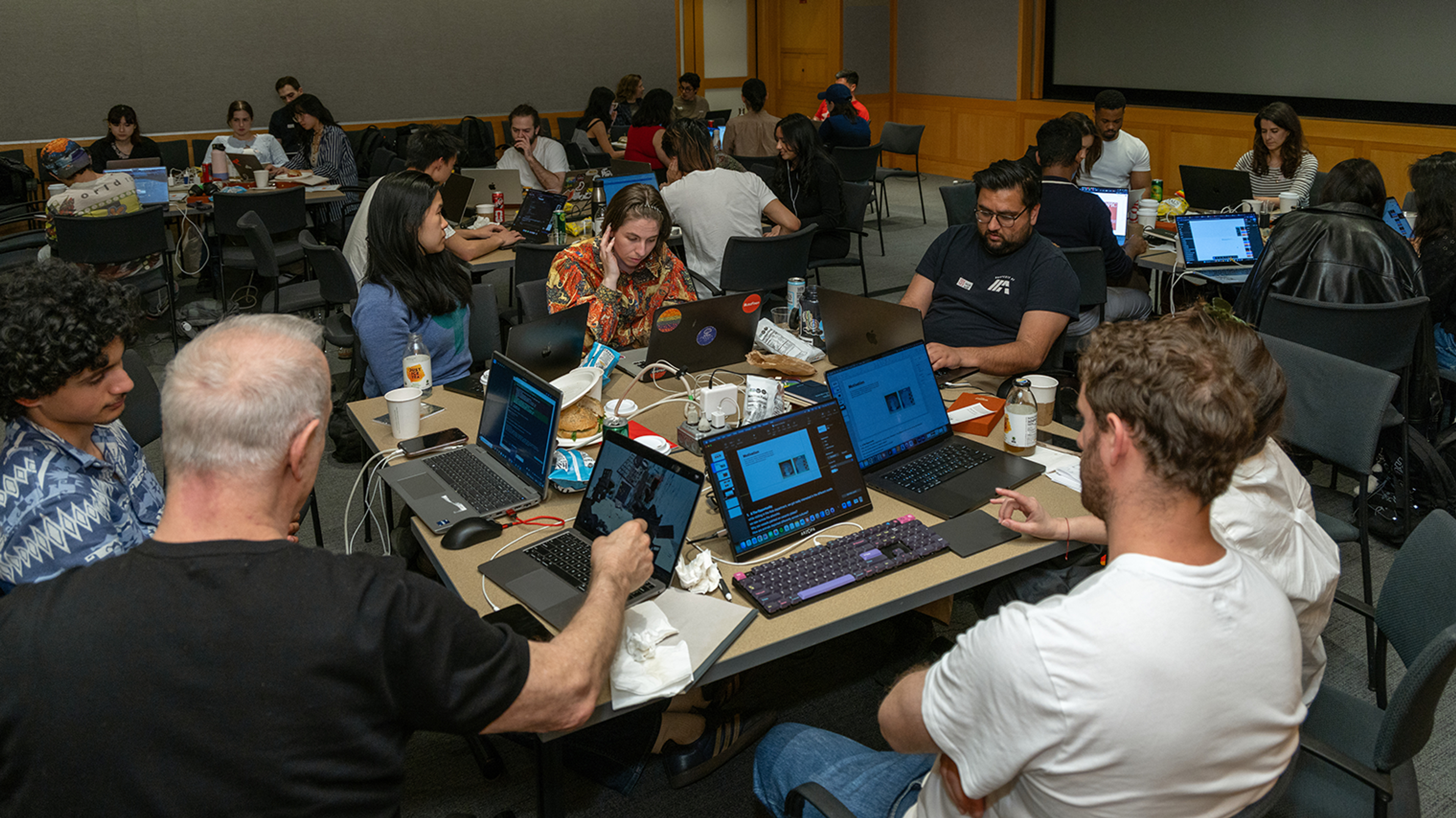 A busy room with diverse people engaged in collaborative work on laptops around tables. 