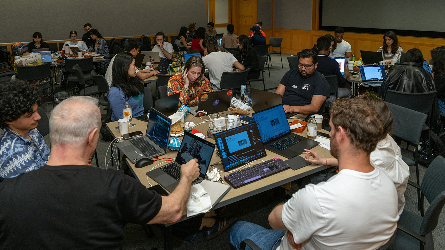 A busy room with diverse people engaged in collaborative work on laptops around tables.