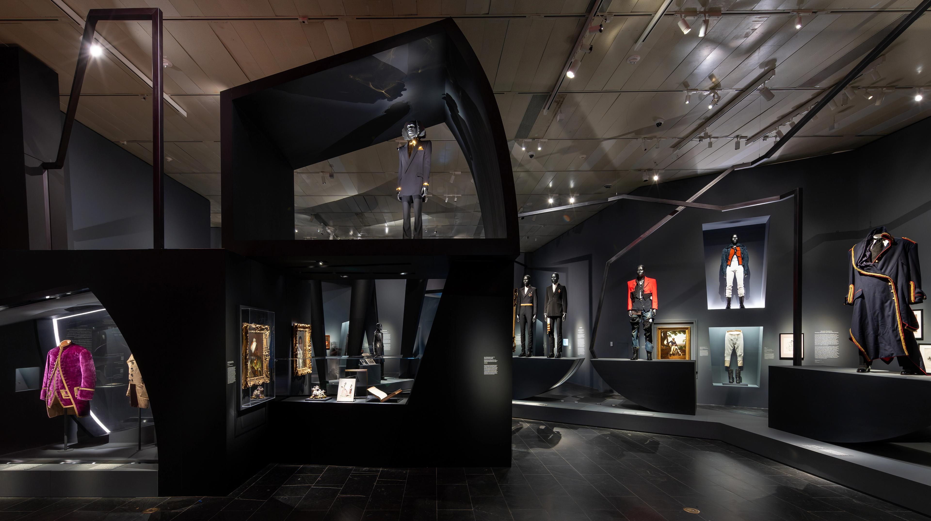 Dark gallery space with both angular and curvy display frames, each spotlighting items of menswear.