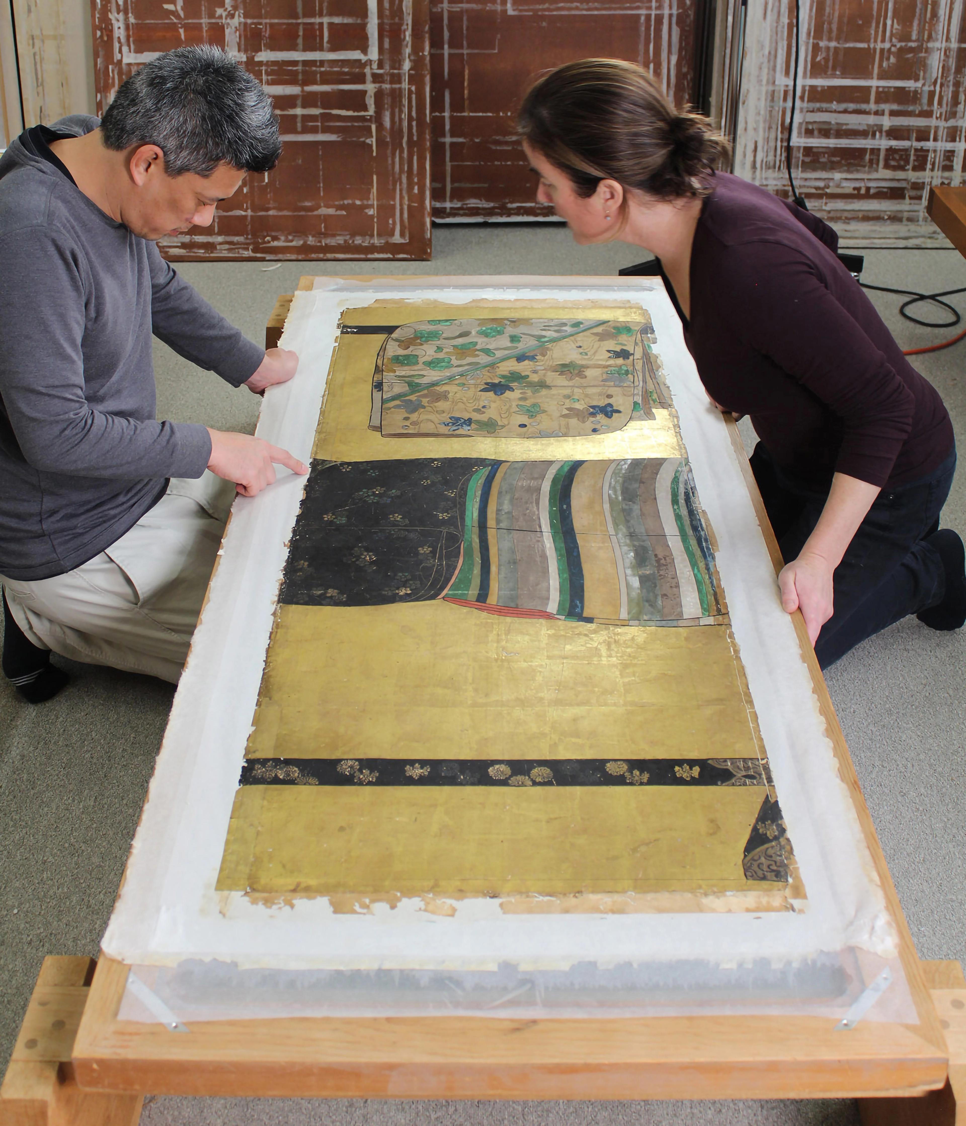 Two conservators kneeling over golden folding screens