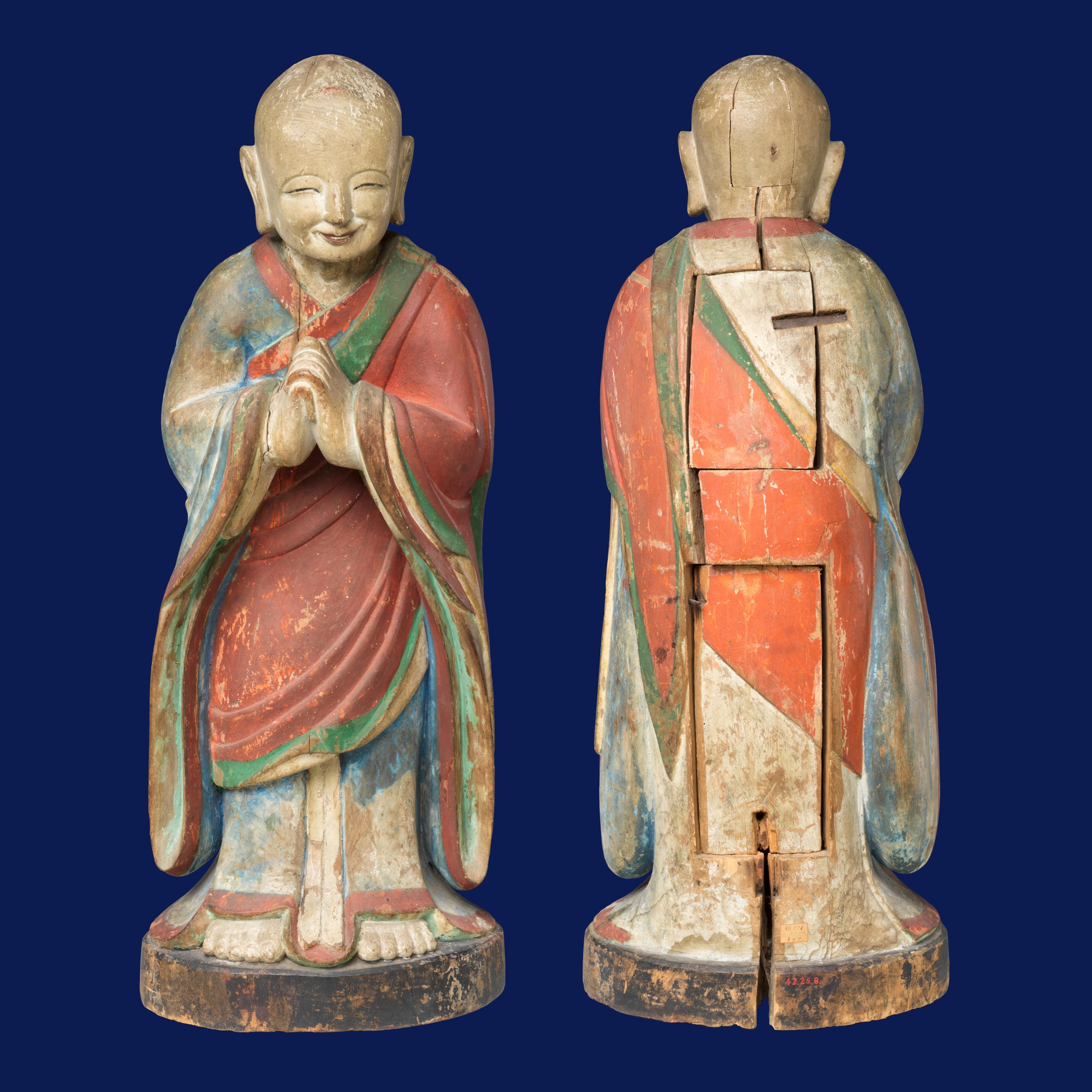 Carved wooden statue of a smiling monk in a red and blue robe, hands together in prayer. Front and back view on a dark blue background.