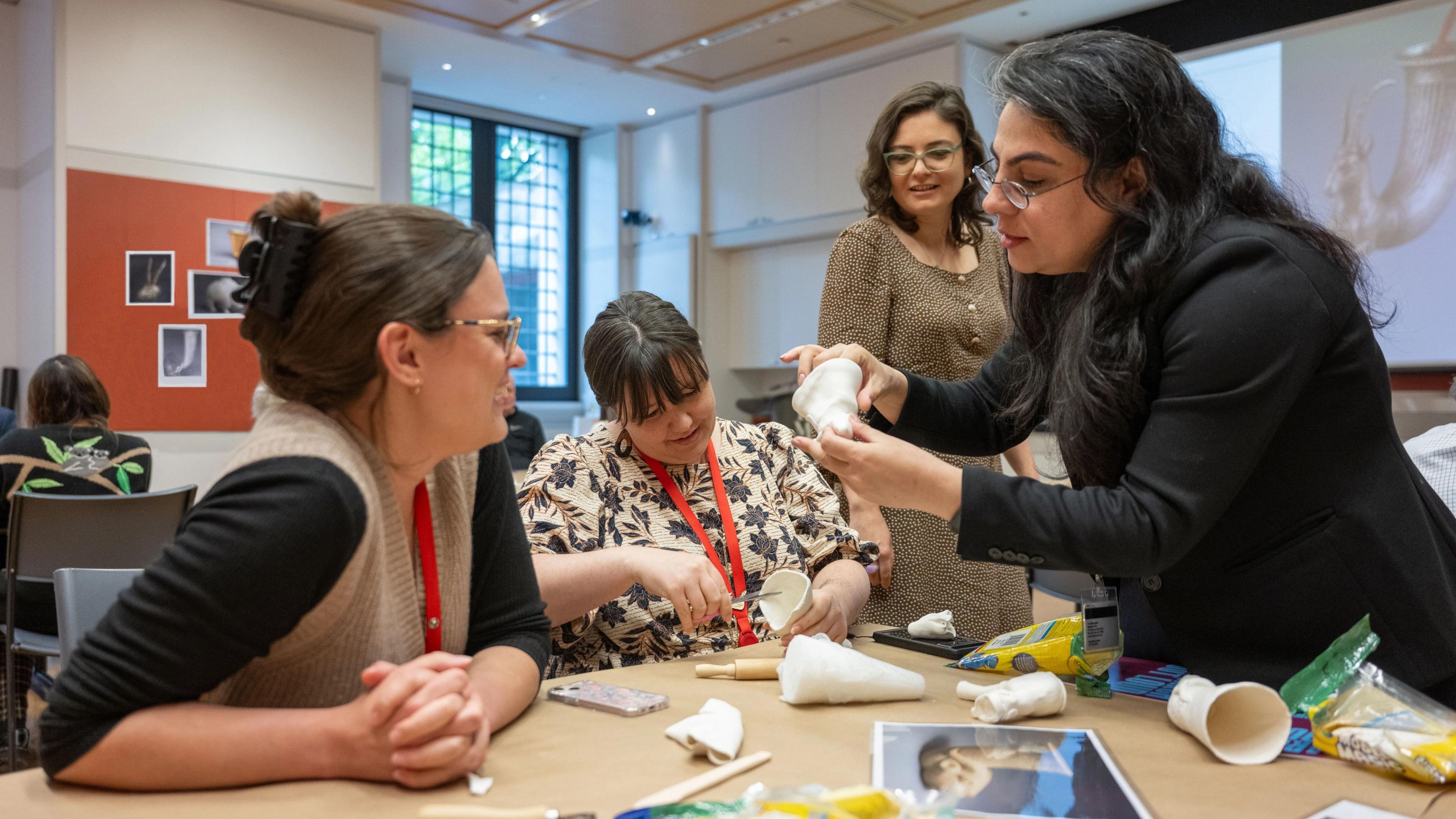 Four women take part in a workshop involving white clay around a table with tools, images, and supplies. One woman trims clay while seated. An instructor beside her demonstrates a technique as the two other women watch. The space is bright and focused, with a pottery image on a projector screen in the background.