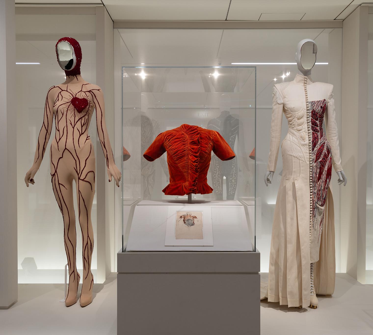 A museum exhibit displays avant-garde fashion. A mannequin in a nude bodysuit with red vein patterns and heart, next to a white gown with red details. A ruched red top is encased.