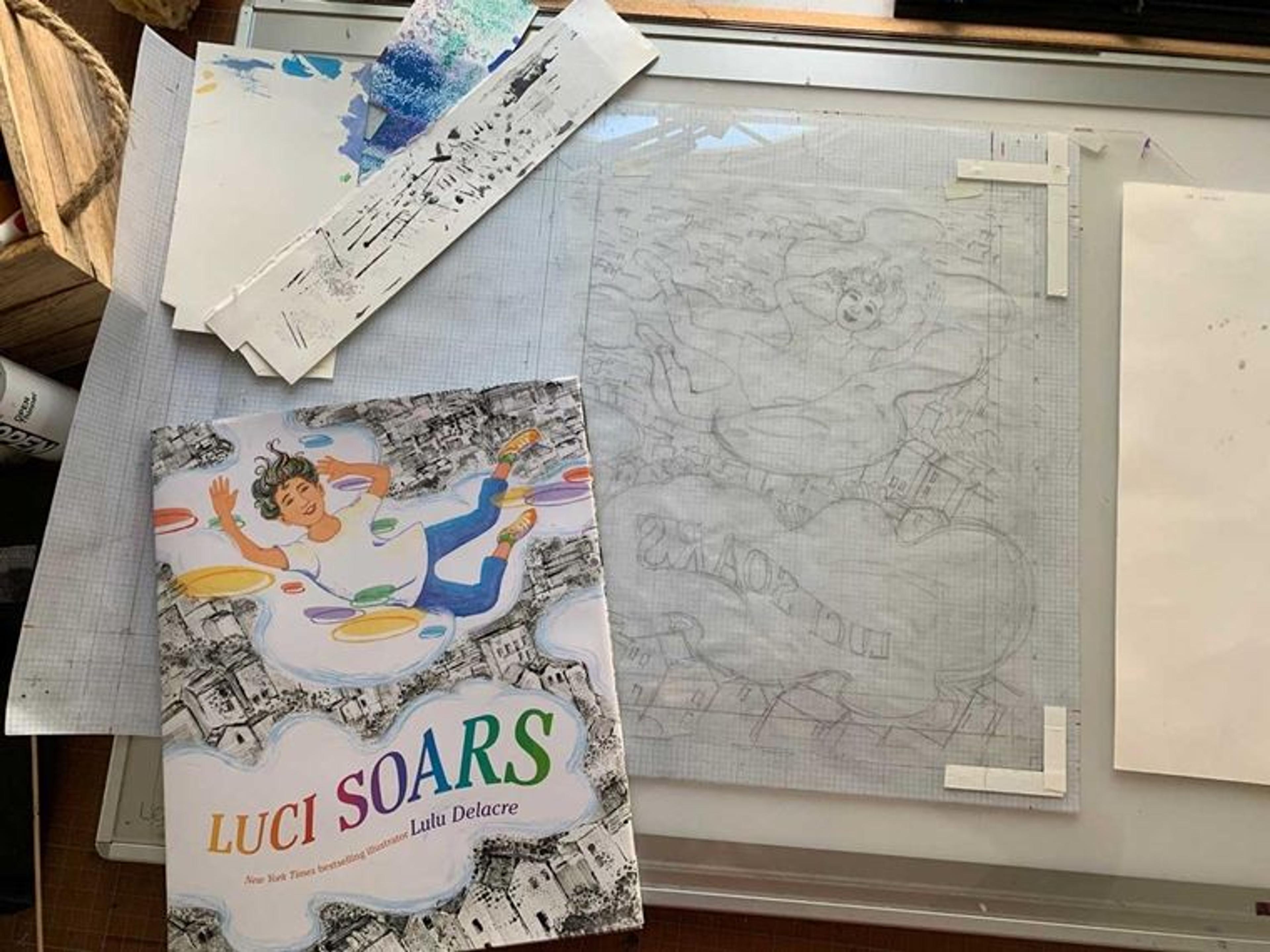 On top of a work bench, a copy of the book Luci Soars is placed atop a rough pencil sketch on tracing paper. The pencil sketch is the mirror image of the book cover.