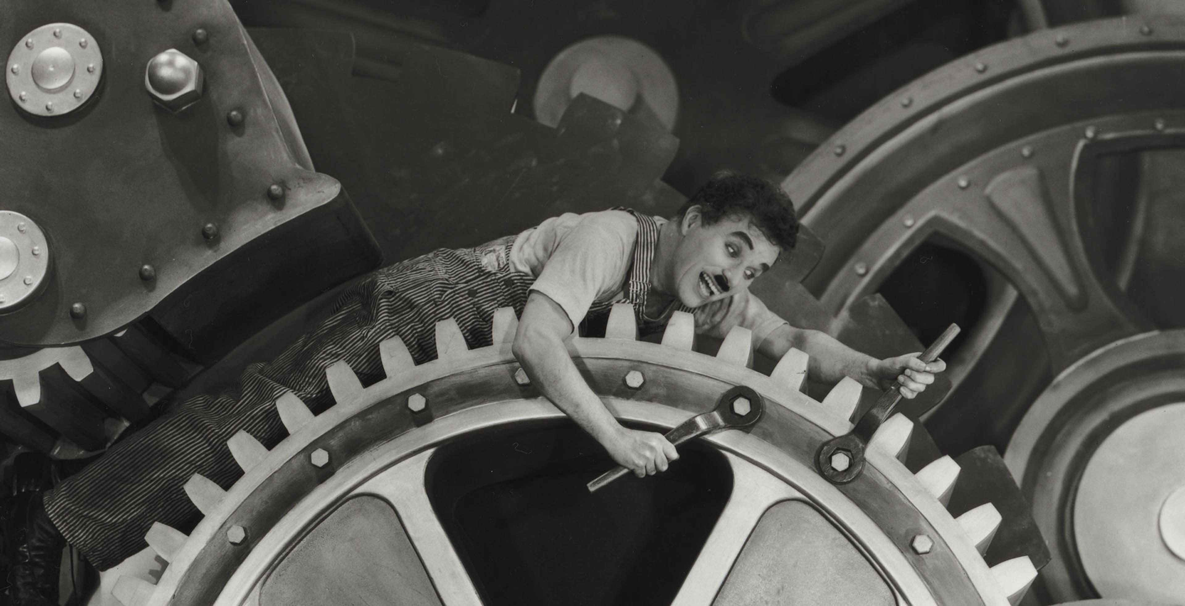 Black and white image of the actor Charlie Chaplin dressed as the tramp, a white man in overalls in this scenario and a mustache where he is trapped between massive factory cogs that he is riding while also trying to tighten the bolts with both of his hands at the same time.