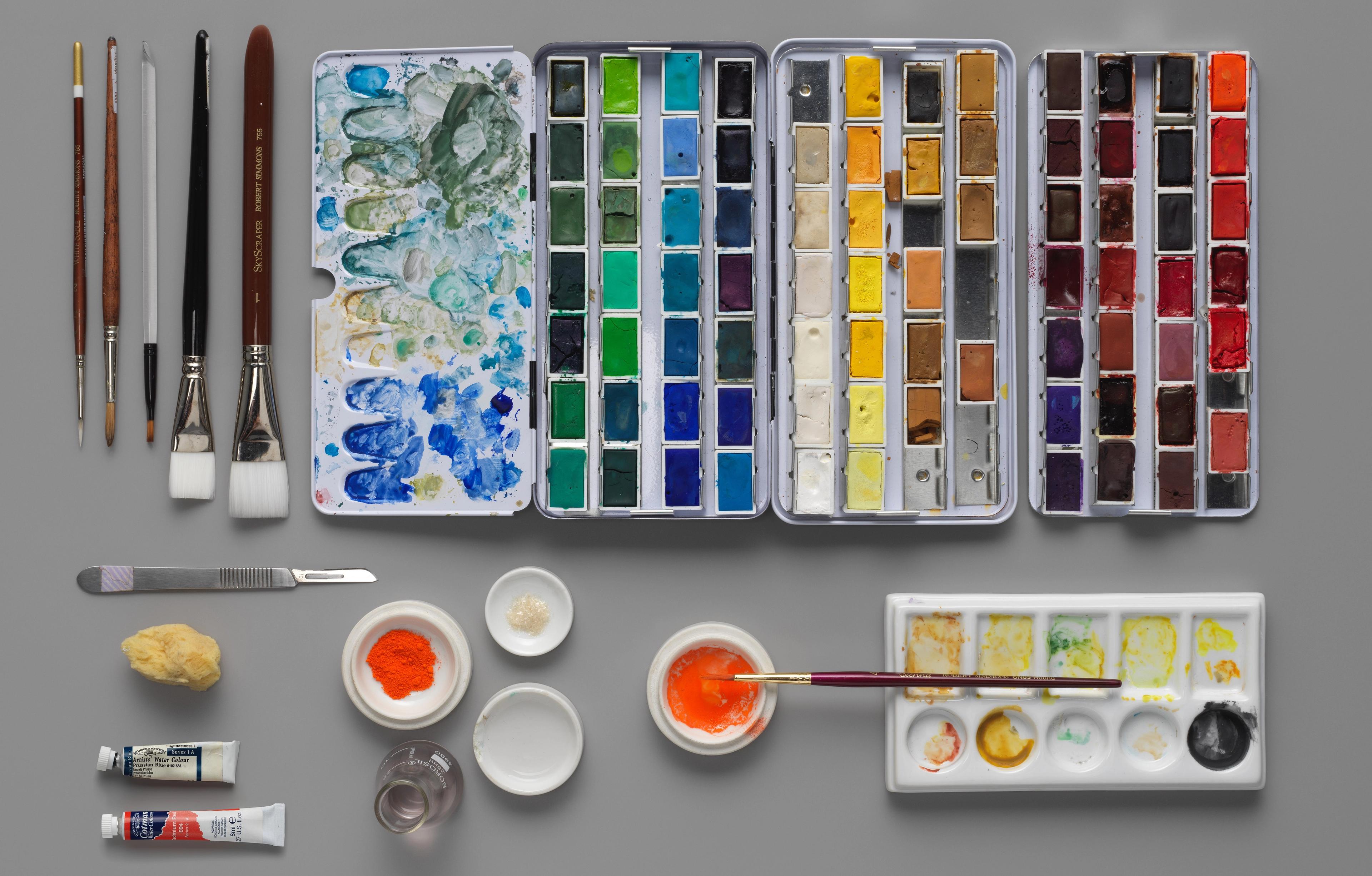 A neat arrangement of brushes, knives, watercolor pans, and other tools on a grey tabletop.