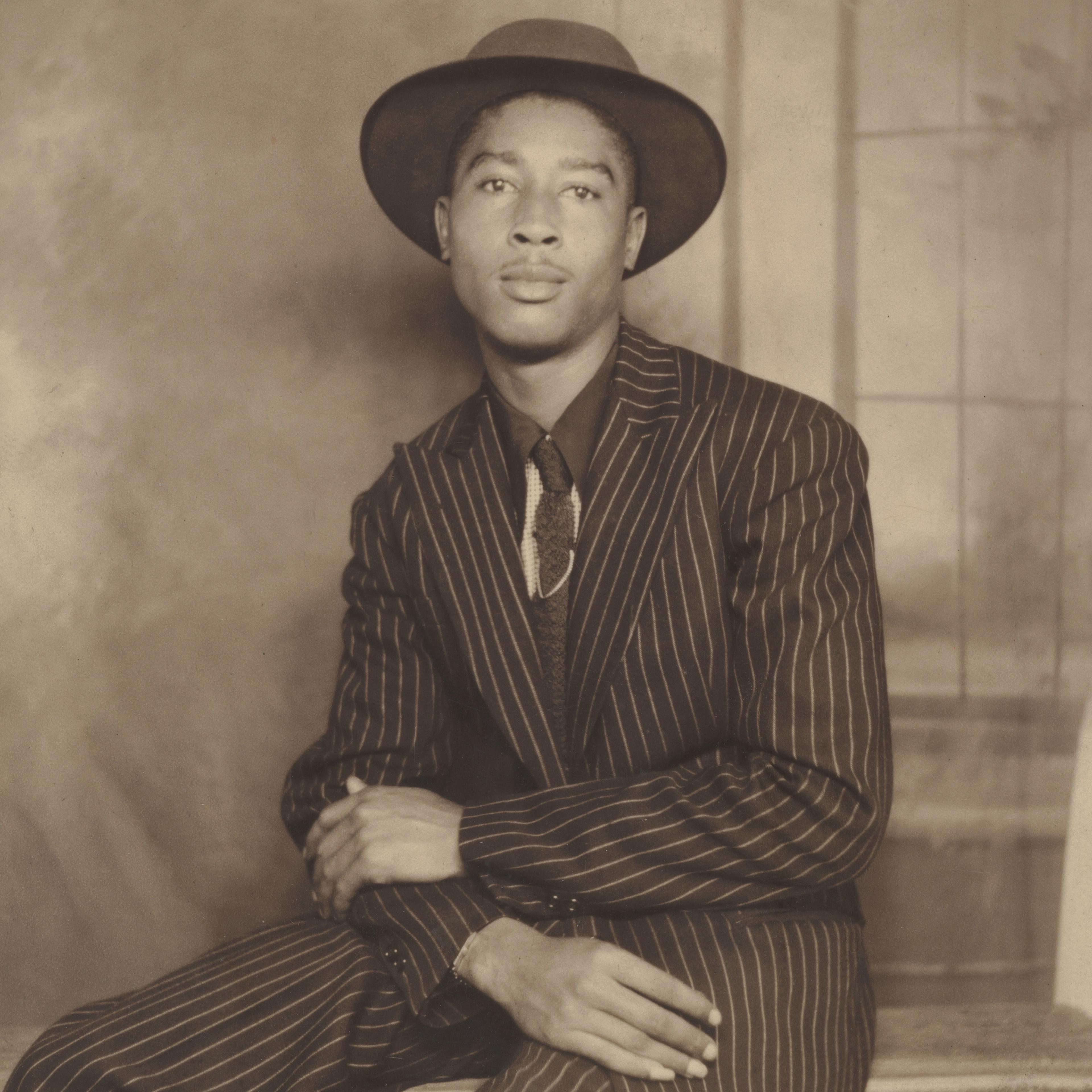 Cropped studio portrait of a well-dressed man in a pinstripe suit, tie, and hat. He is seated, leaning slightly to the right with his arms crossed in his lap.