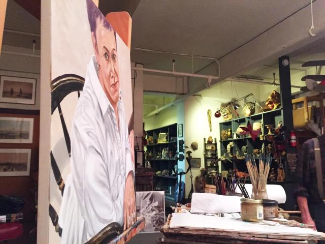 Photograph of a busy artist's studio. A portrait and table are in the foreground, while shelves of full of objects line the walls in the background.