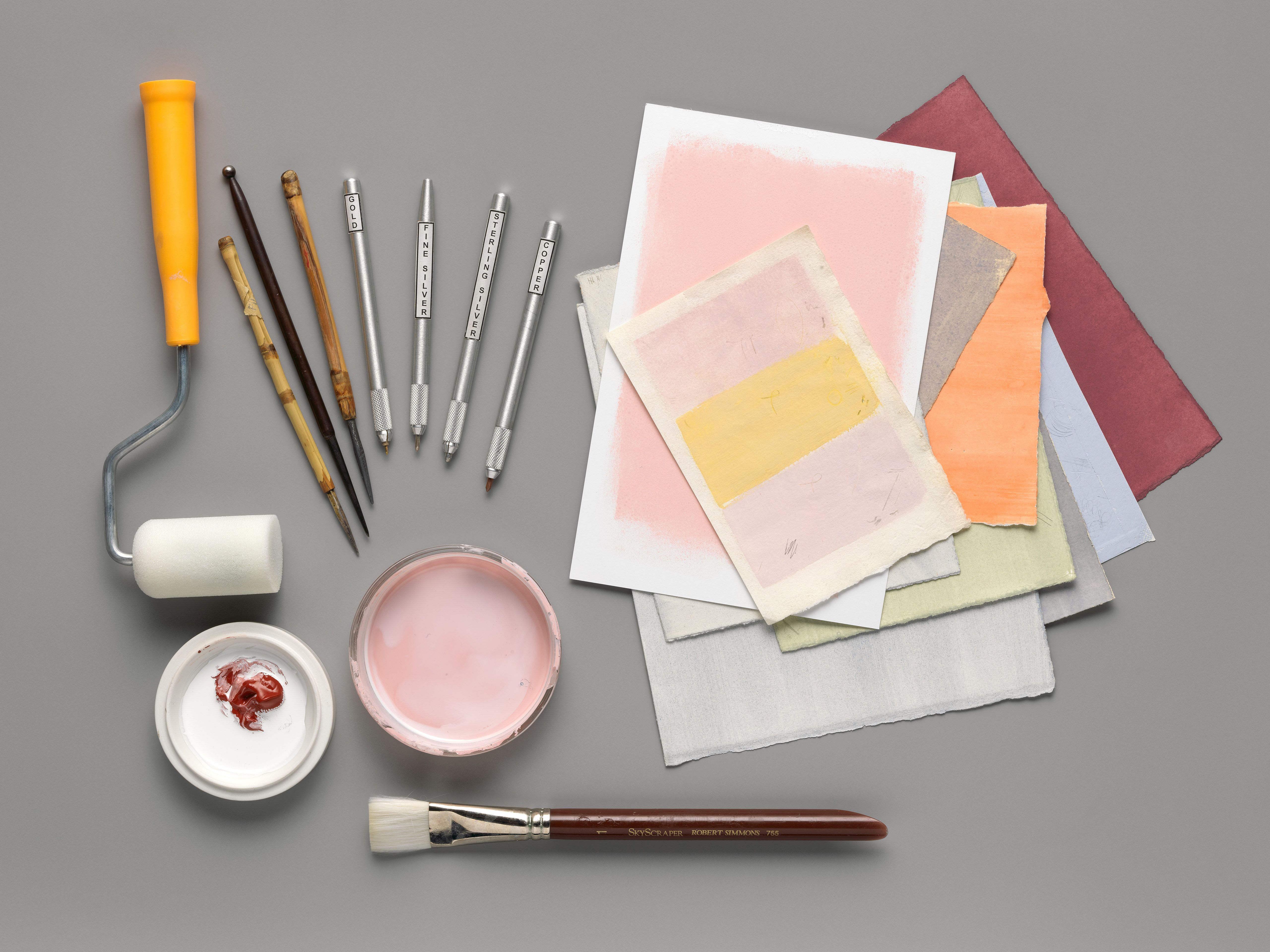 An artful arrangement of specialized tools for making metalpoint drawings beside a stack of colorful paper.