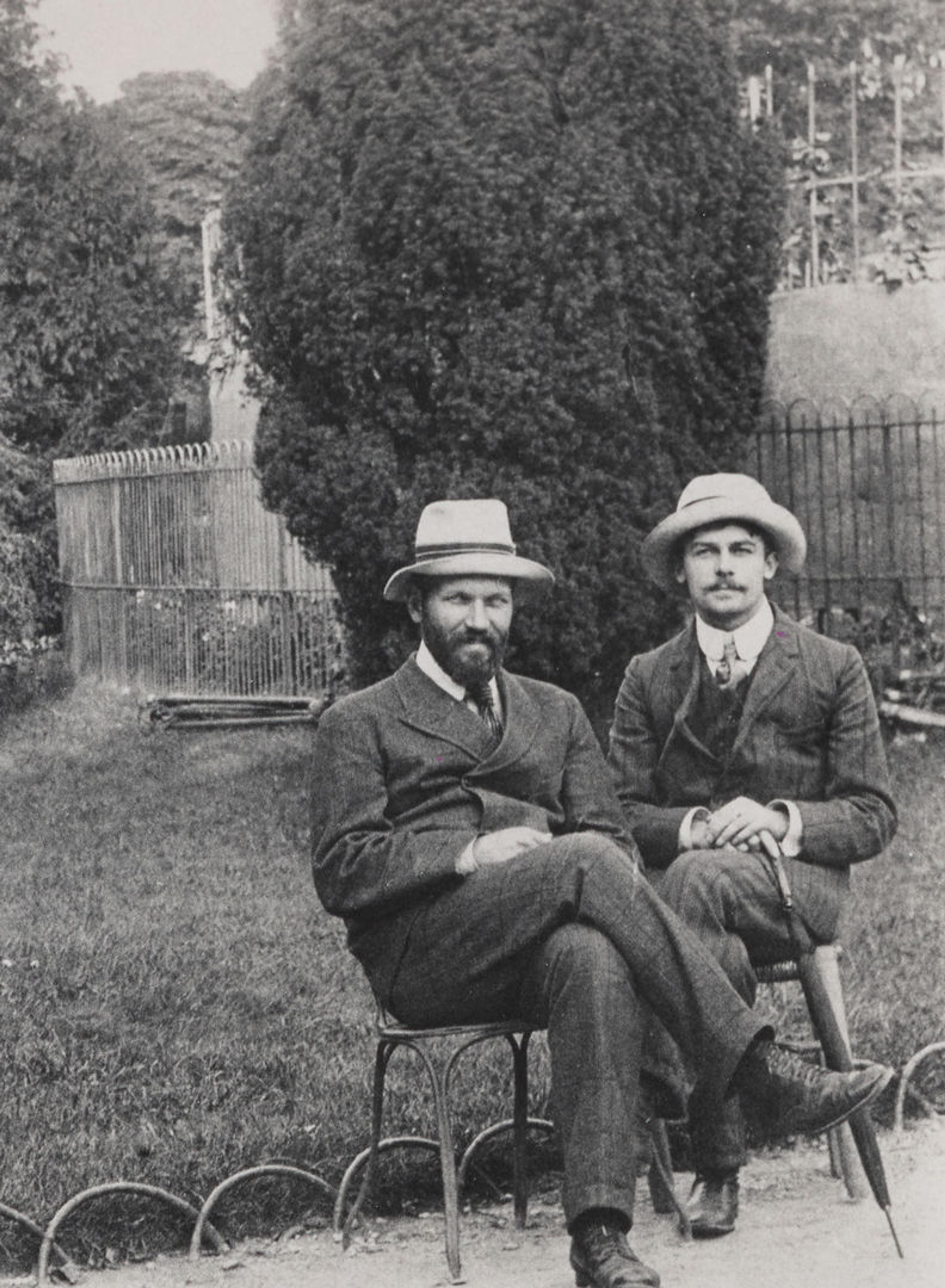 Black and white image of two white men in suits wearing hats in front of a lawn and large bush in the background.