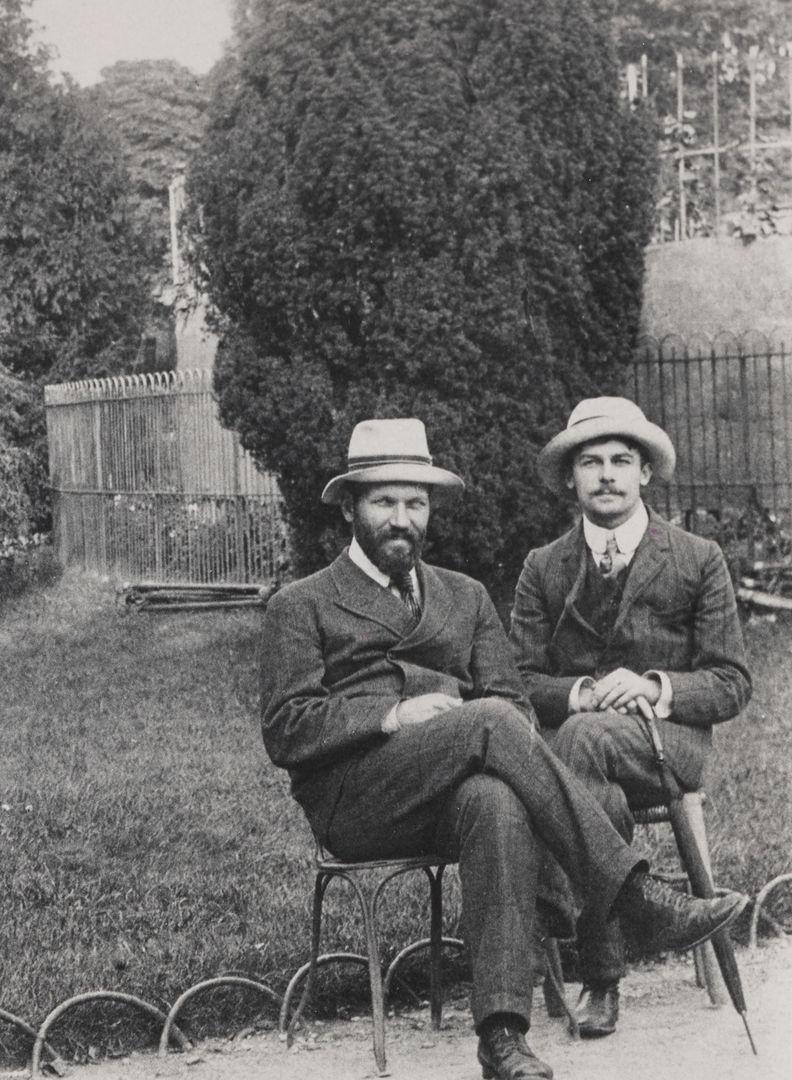 Black and white image of two white men in suits wearing hats in front of a lawn and large bush in the background.