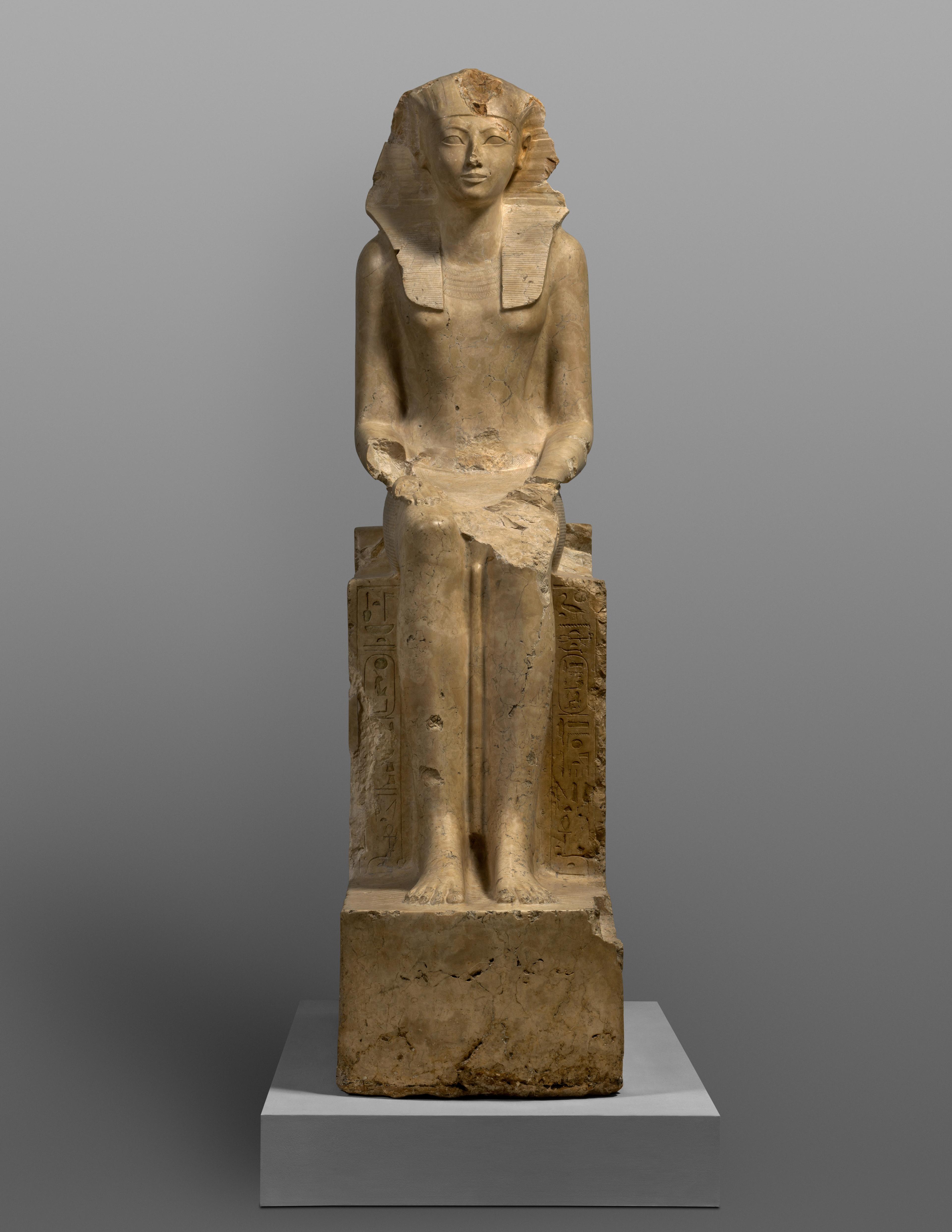 Ancient Egyptian limestone statue of a seated Hatshepsut. The statue shows damage to her arms, knee, and head piece