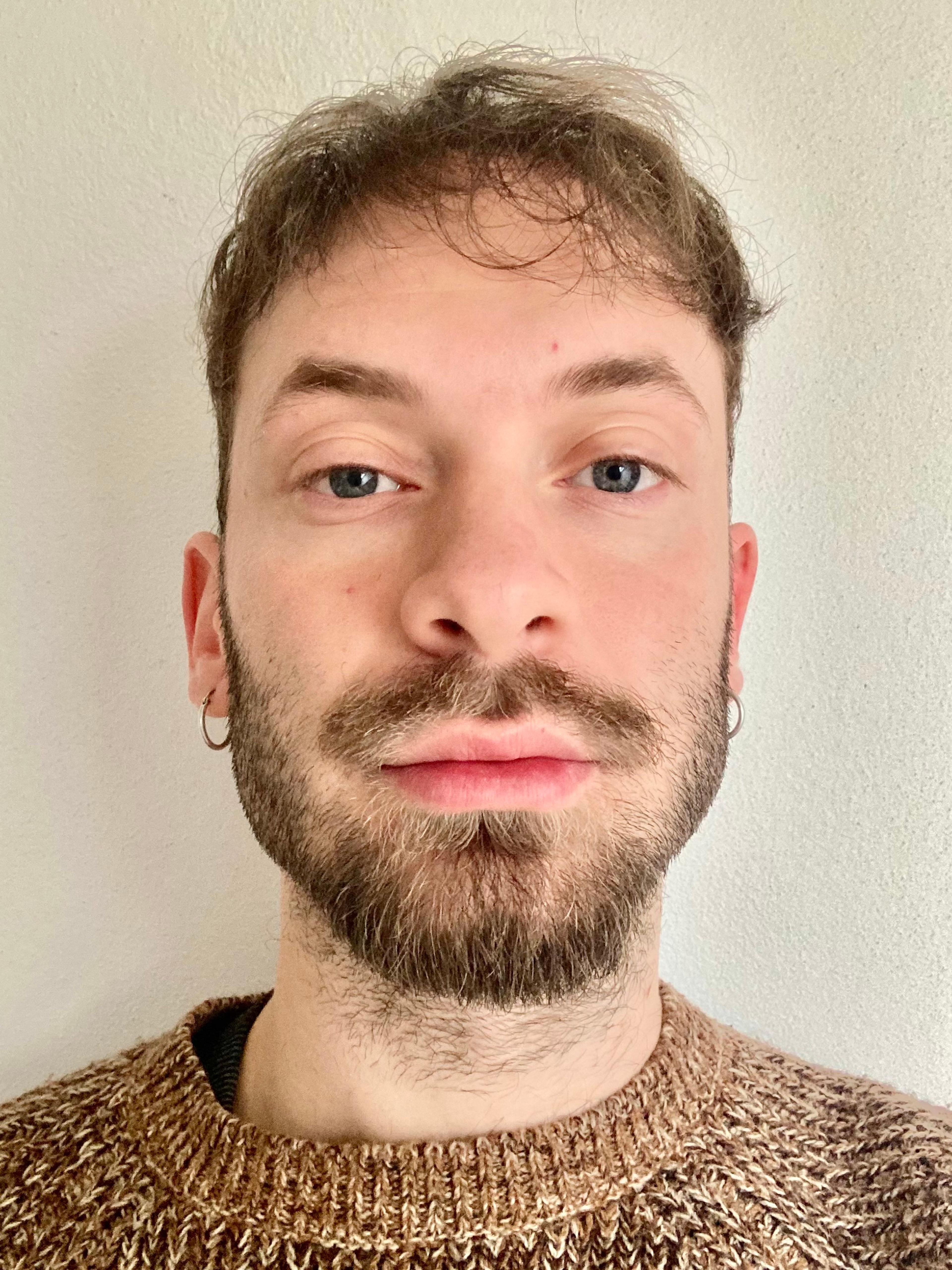 A man with short hair and a beard poses in front of a white wall