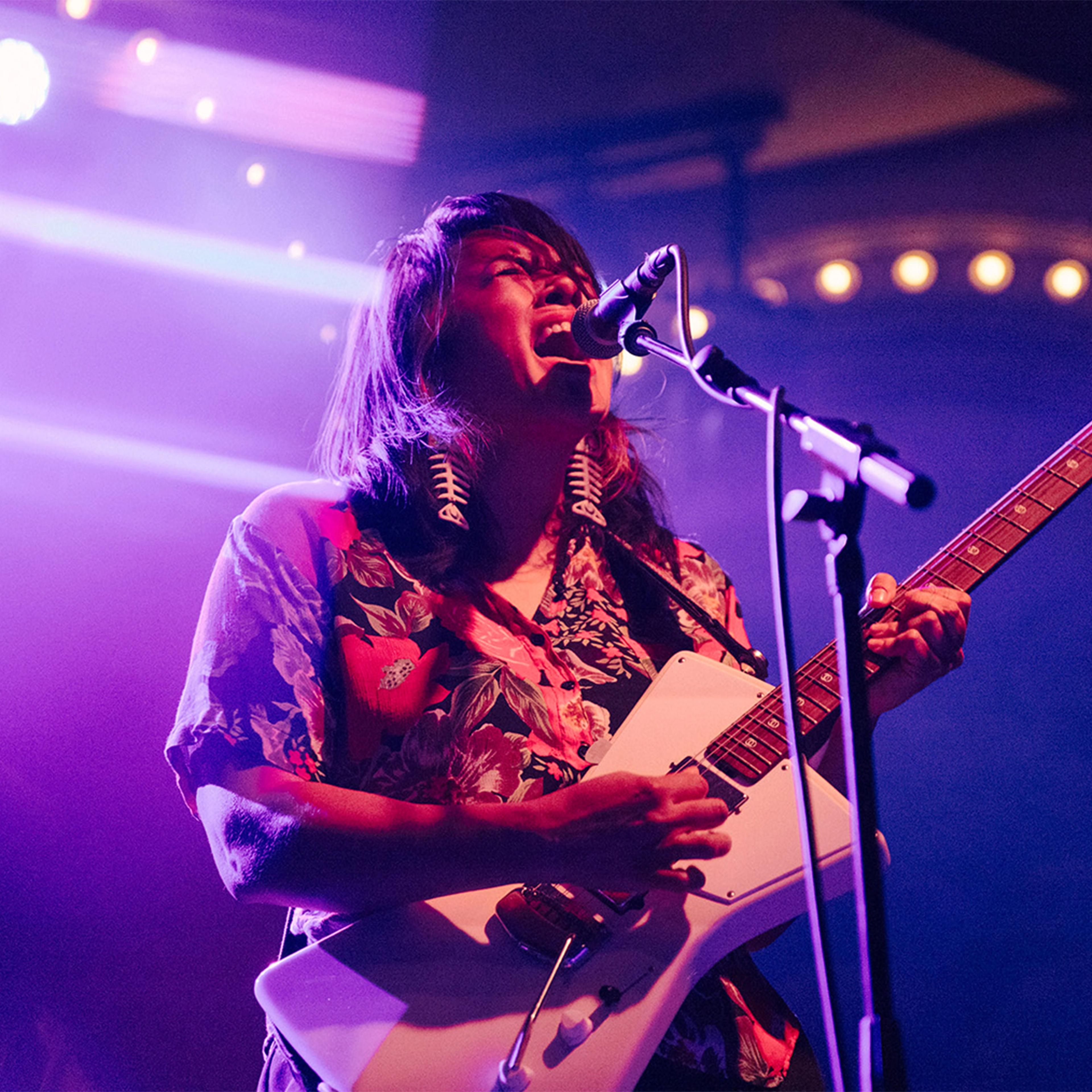 A long-haired musician sings passionately into a microphone while playing a white electric guitar onstage, bathed in vivid purple and red lights.