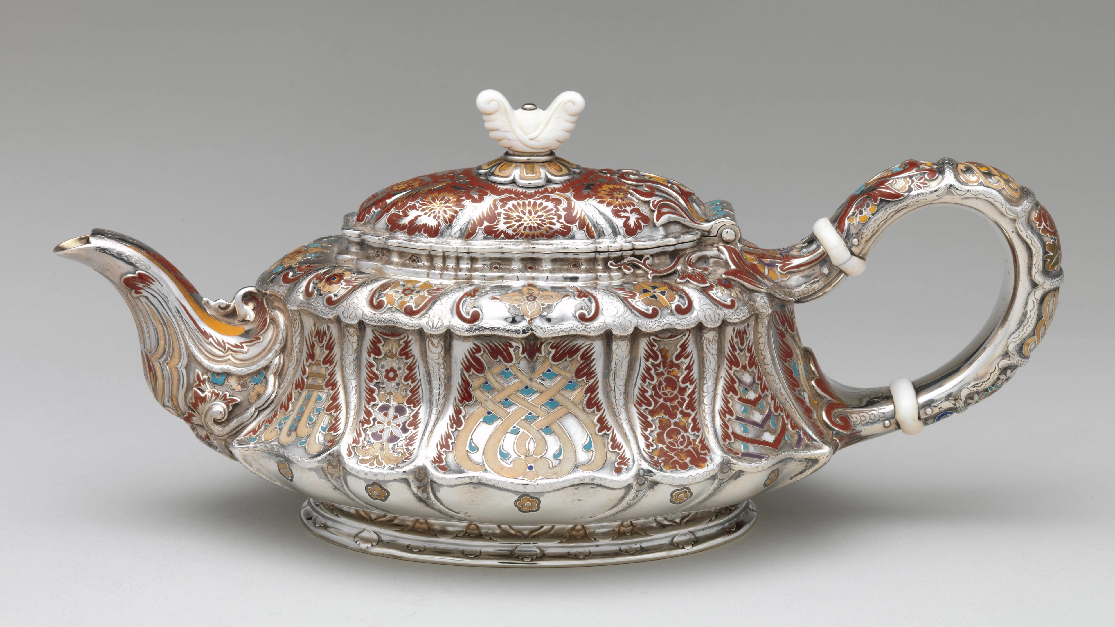 A short but elongated silver teapot with an ivory lid finial adorned with abstract floral motifs rendered in maroon, orange, taupe, and turquoise enamel.