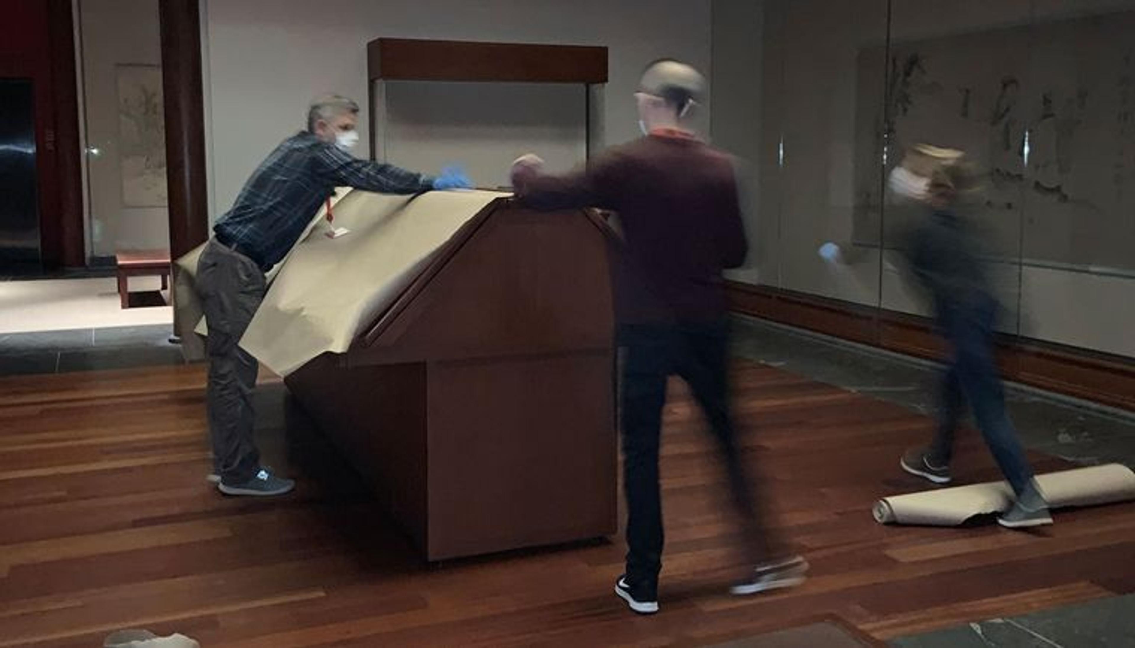 Three people wearing gloves and face masks set butcher paper over a case in the middle of an art museum gallery