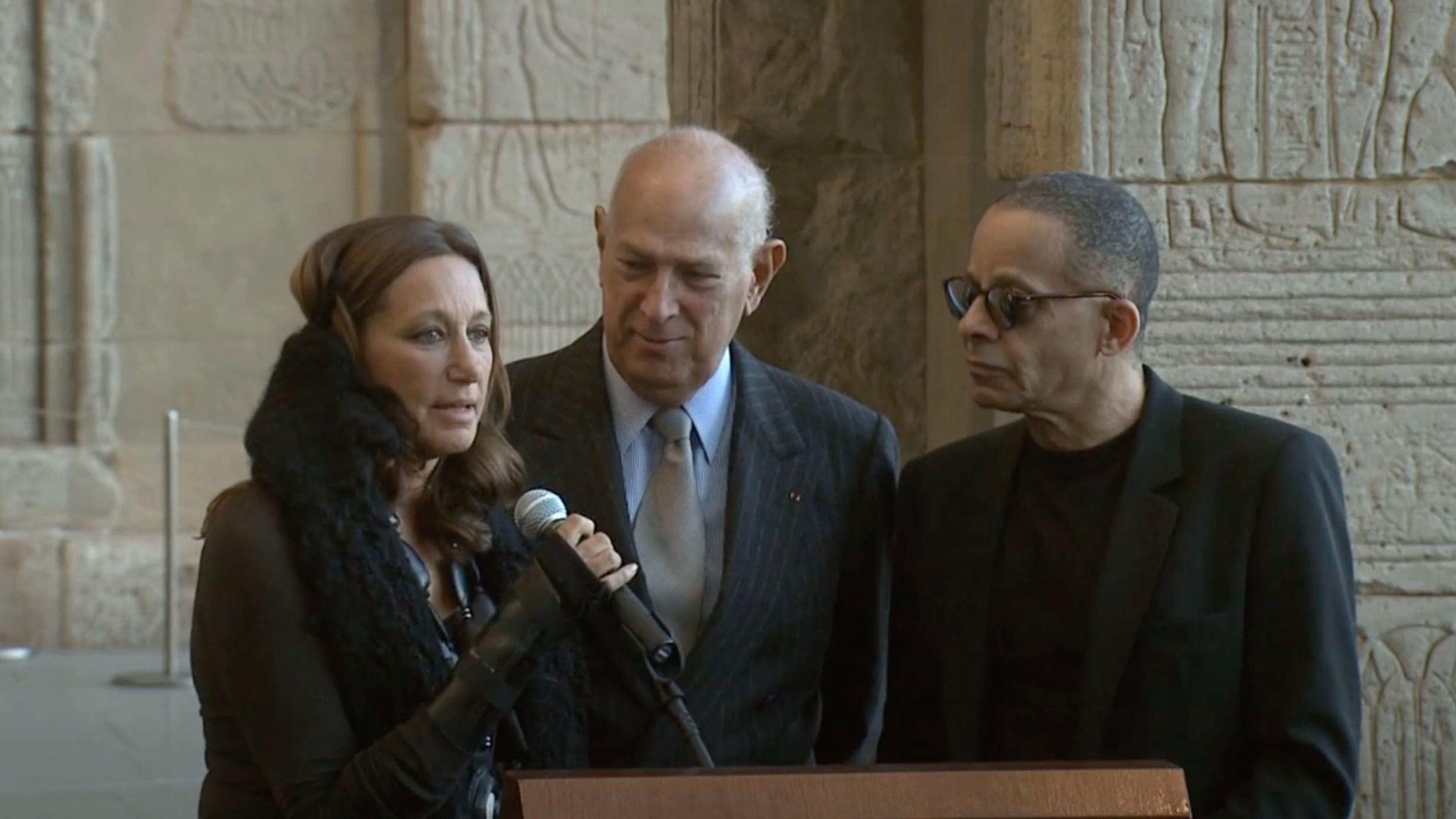 Three people stand at a podium on stage while the woman on the left speaks into a microphone.
