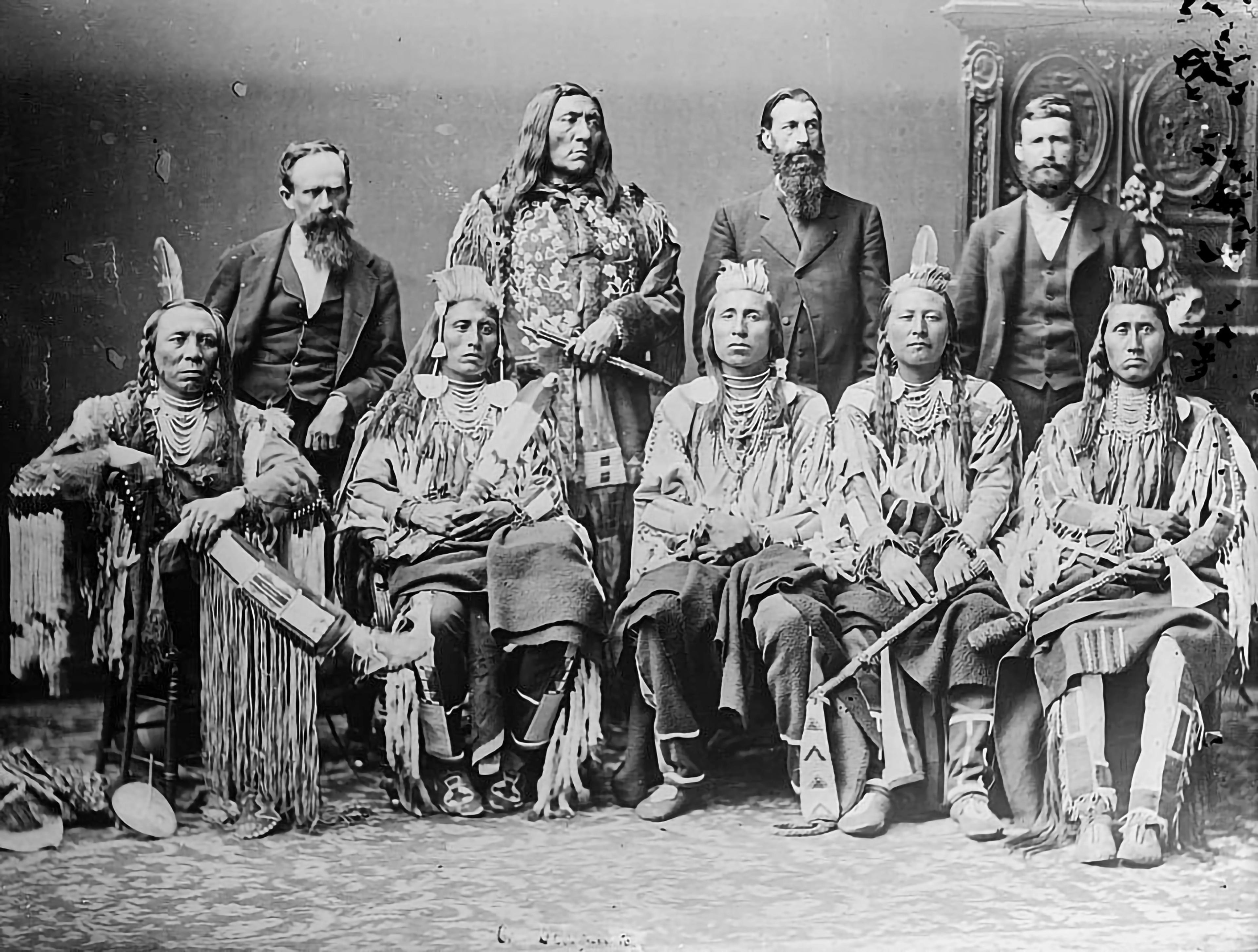 Dated black-and-white photo of several Indigenous people posed with three white men.