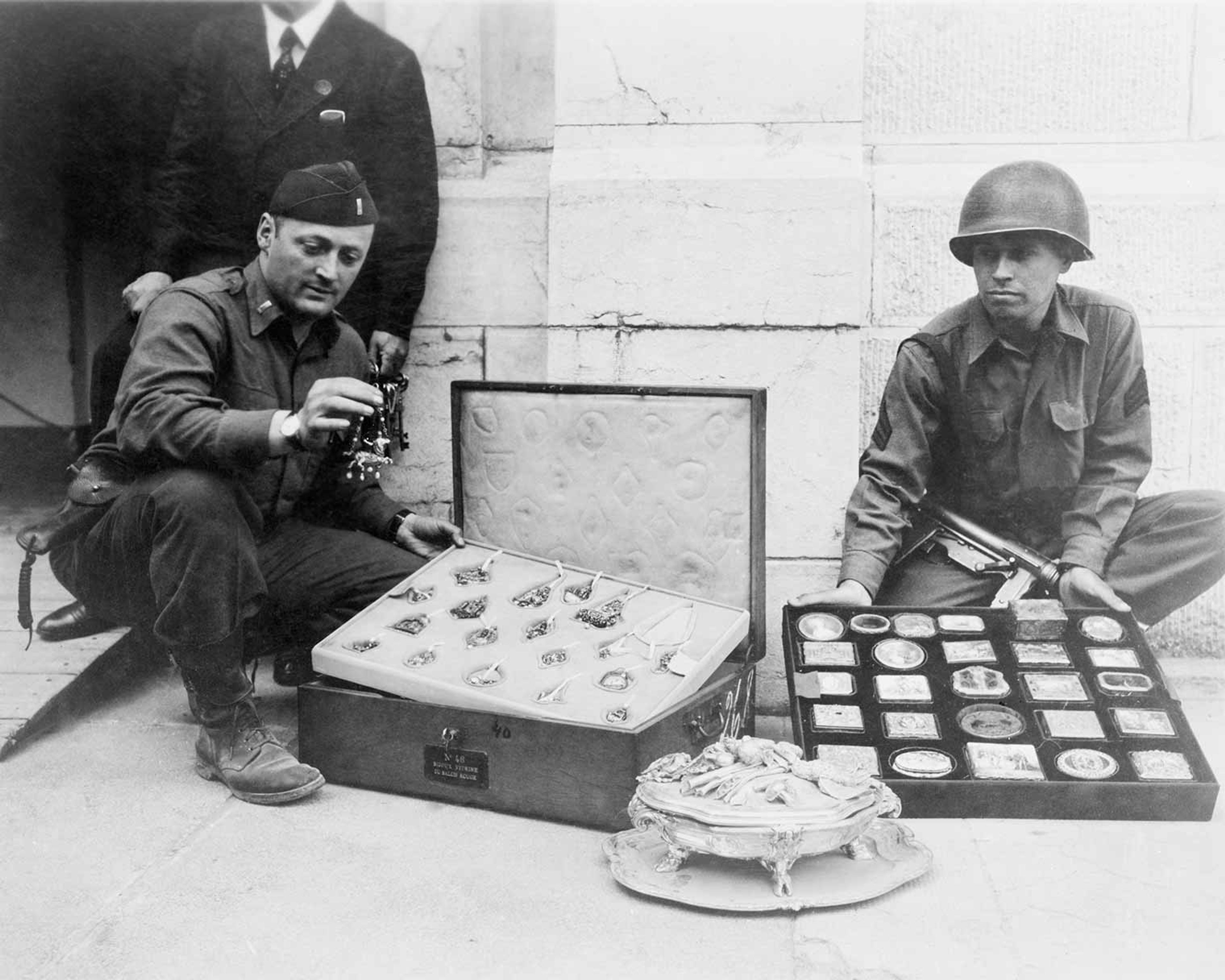 Black and white phot of two soldiers looking at a suitcase with art objects in it