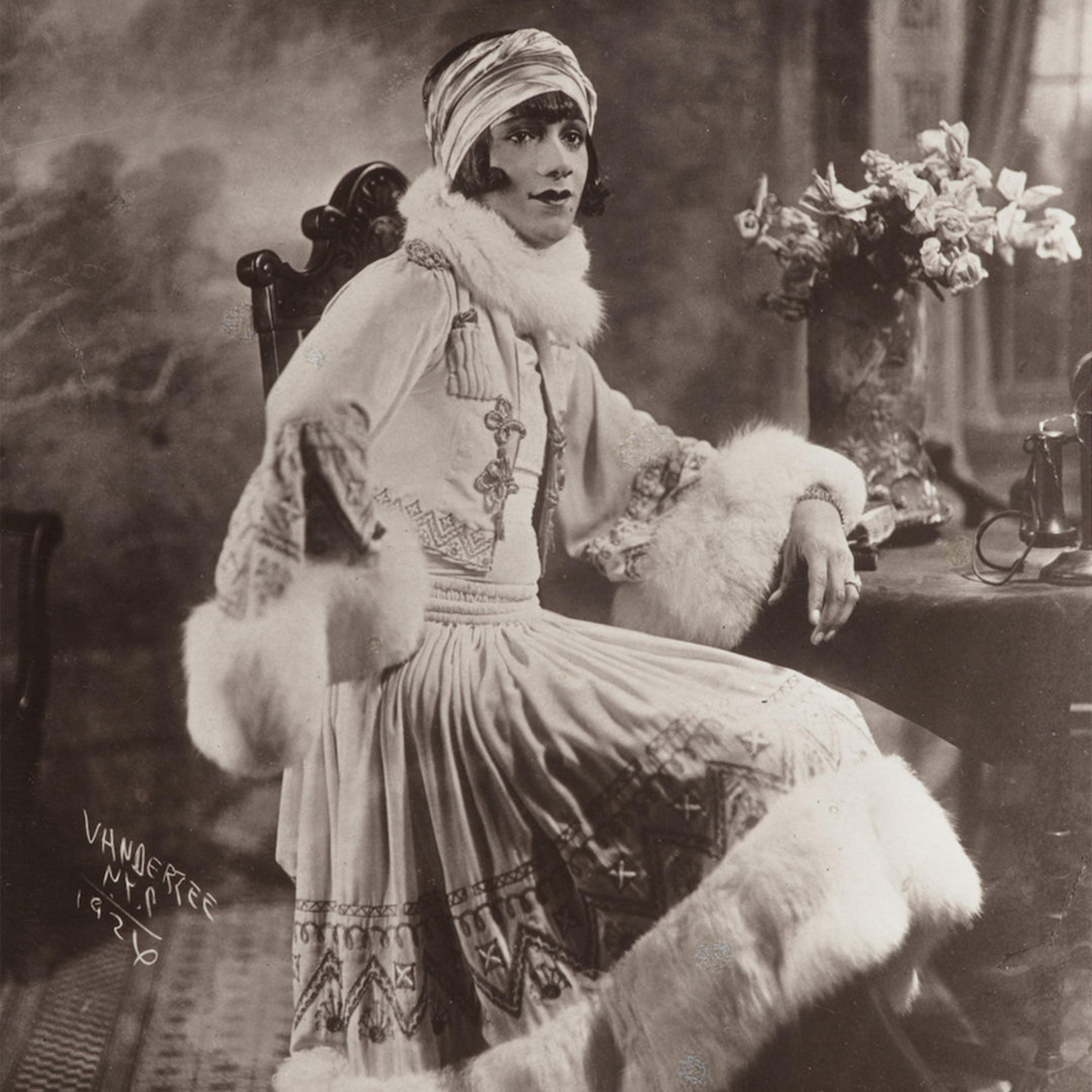 Portrait of woman dressed in heavy garment and fur. Her expression is calm, while her arm rests on the wooden table. 