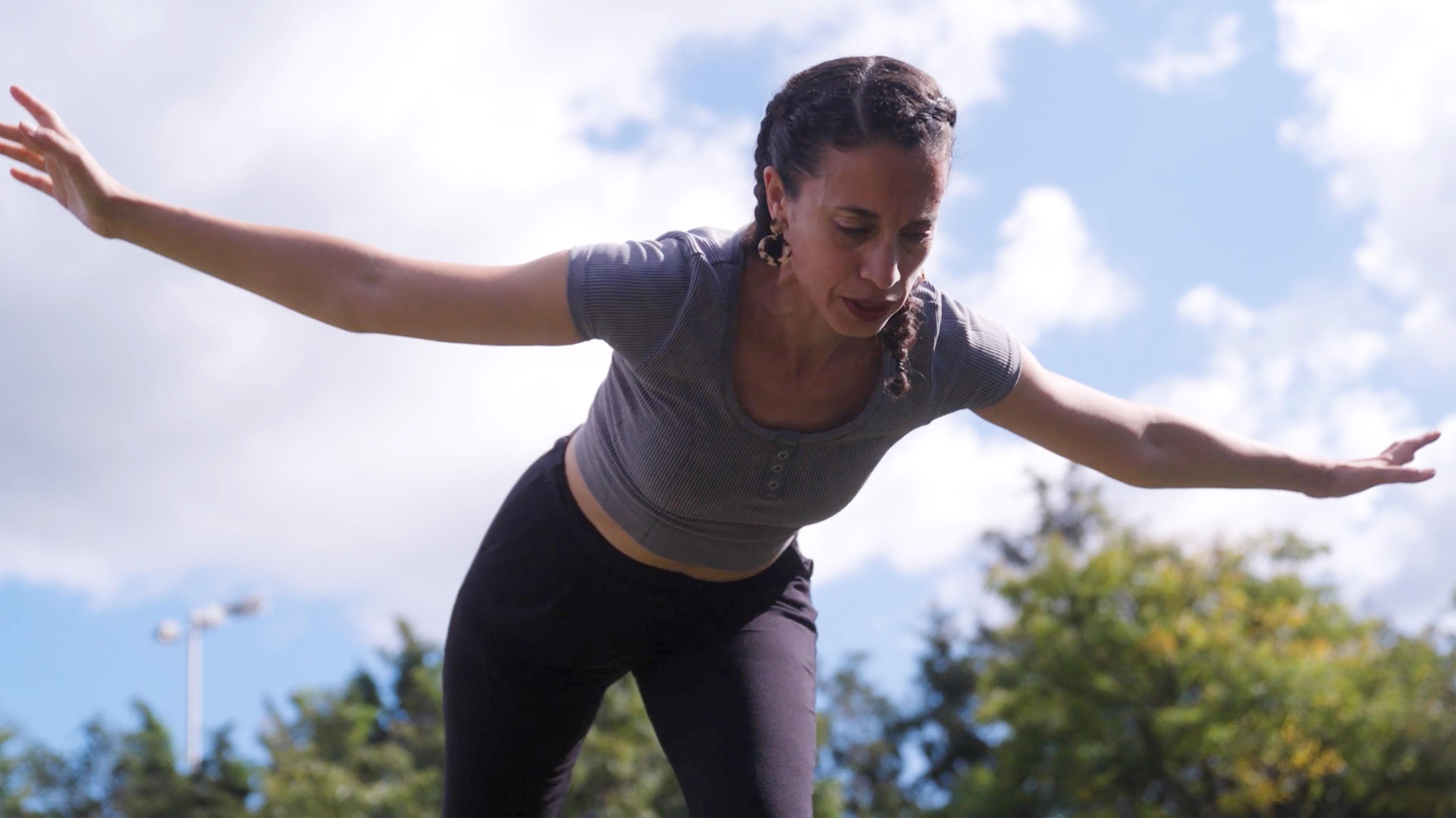 Alethea Pace bends downwards, with both arms outstretched against a cloudy, blue sky