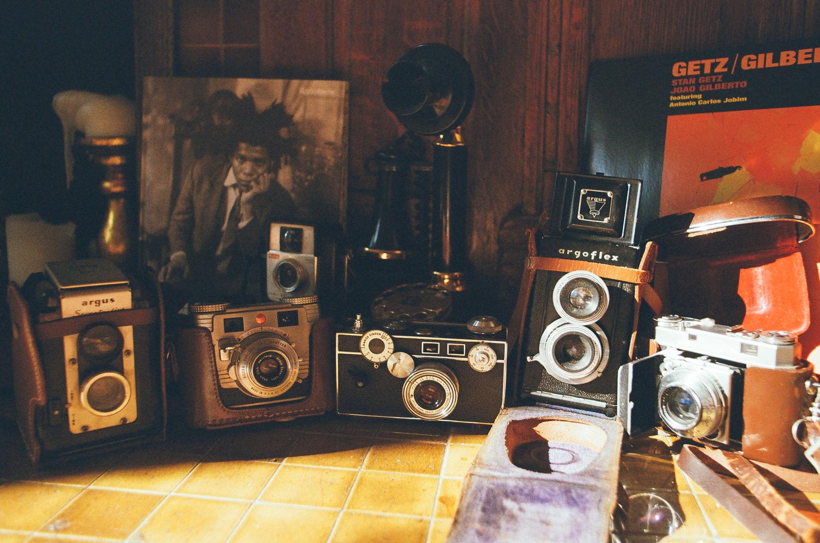 Vintage cameras lined up in a row glint in the sunlight in front of a wooden fireplace mantel