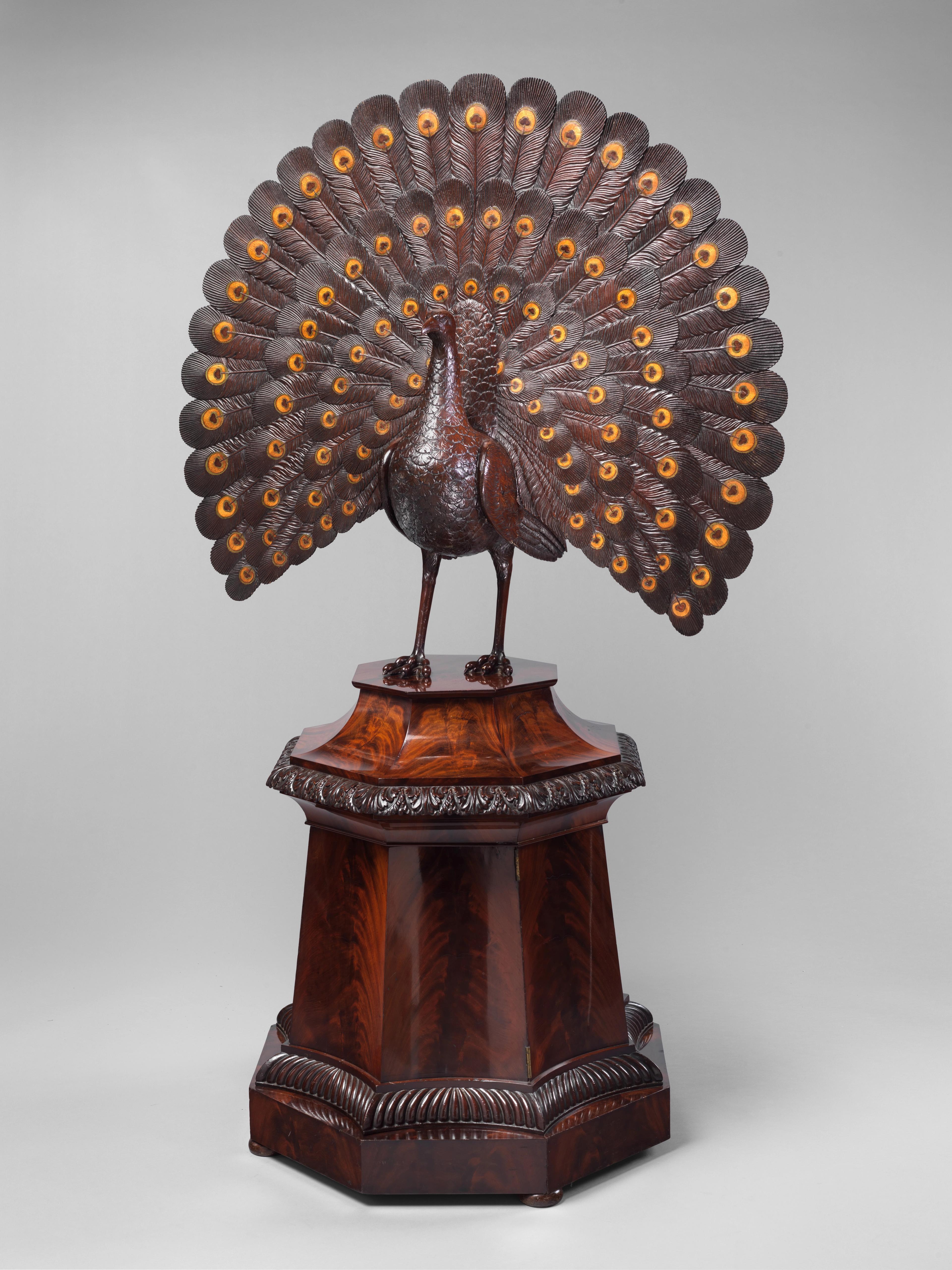 Wooden sculpture of a peacock with an elaborate, fully fanned tail perched atop a glossy wooden pedestal.