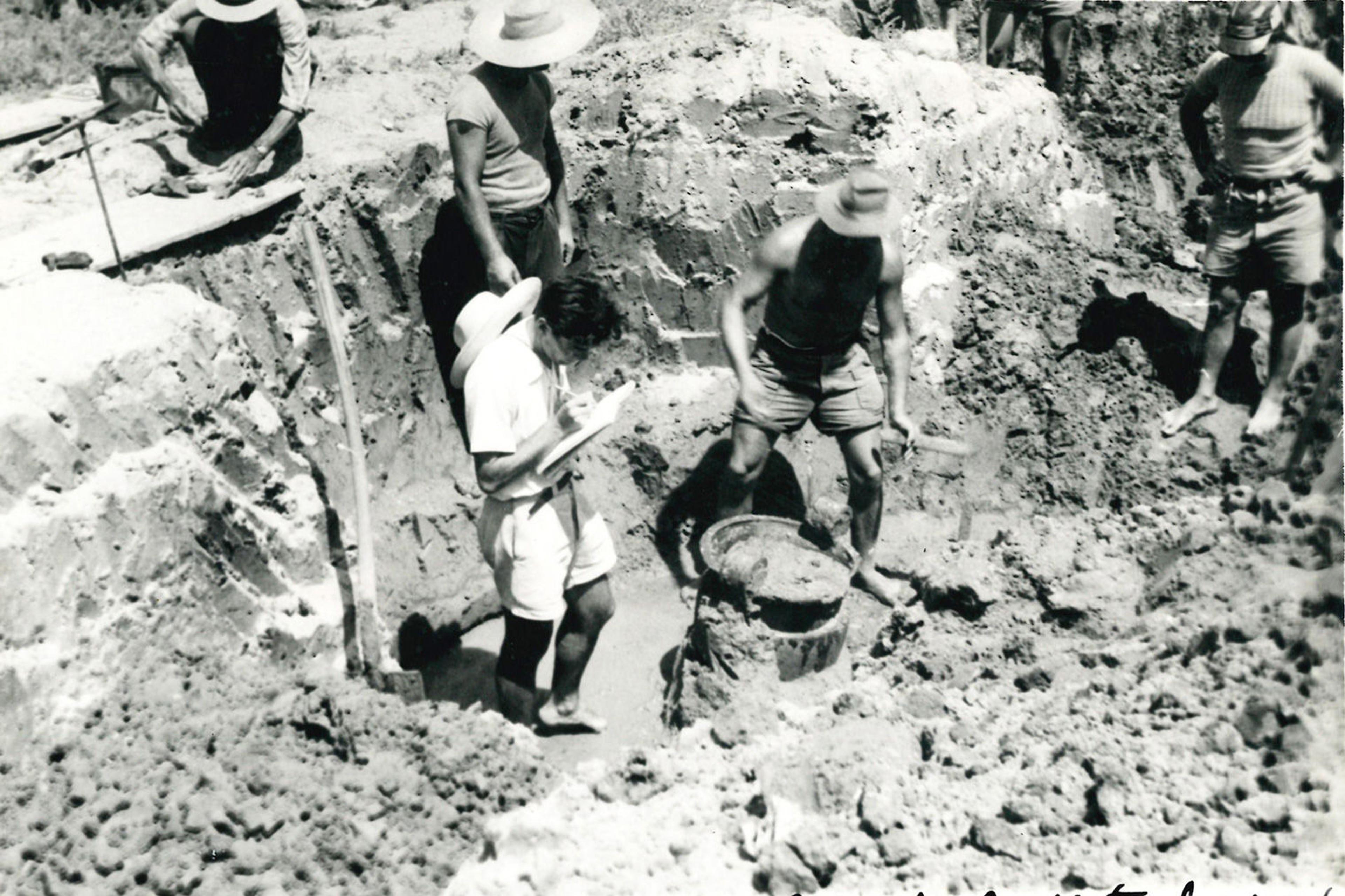 Black and white photograph showing the excavations at Spina. Five figures are at work, and the partially excavated krater sits at the center of the image.