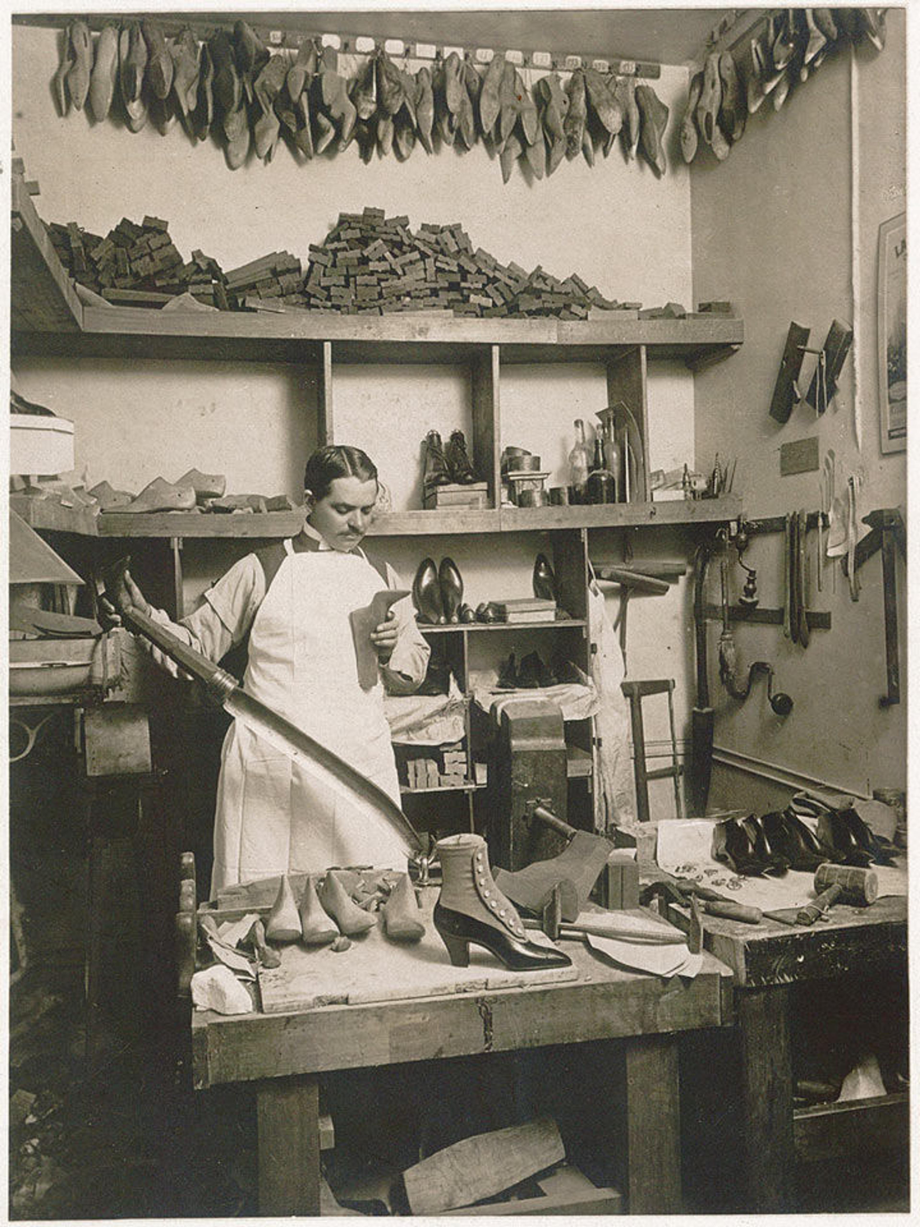 A middle-aged man in a shoemaker's studio holding a last in one hand and the handle of a knife used for rough cutting lasts