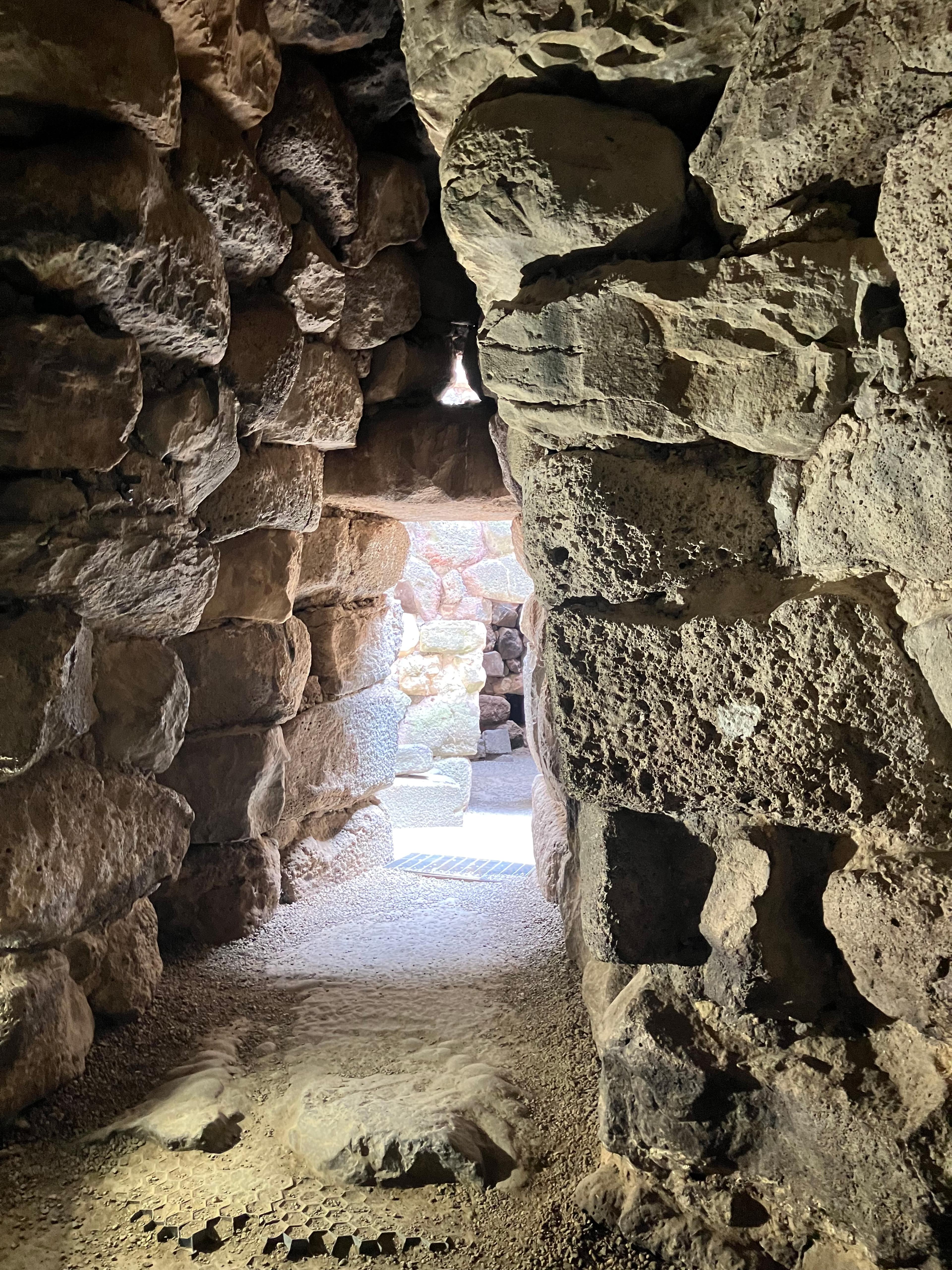 Triangular passageway made of thick rectangular stones, with sunlight pouring in from the far end of the tunnel.