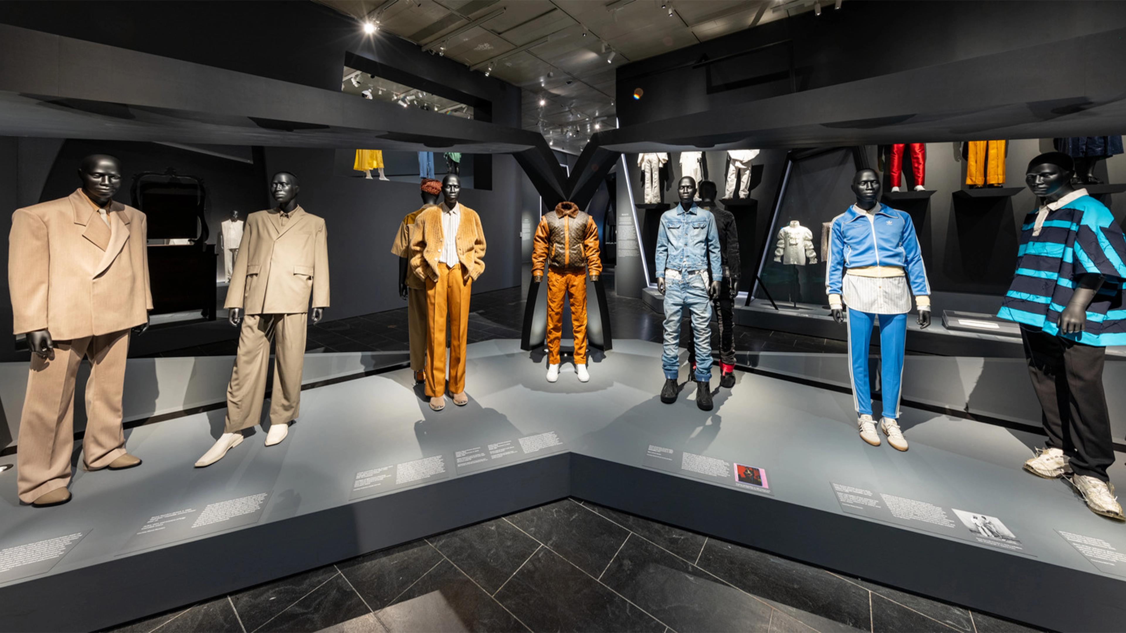 Mannequins in stylish outfits are displayed on a geometric platform in a modern fashion exhibit. The scene is brightly lit, highlighting diverse clothing styles.