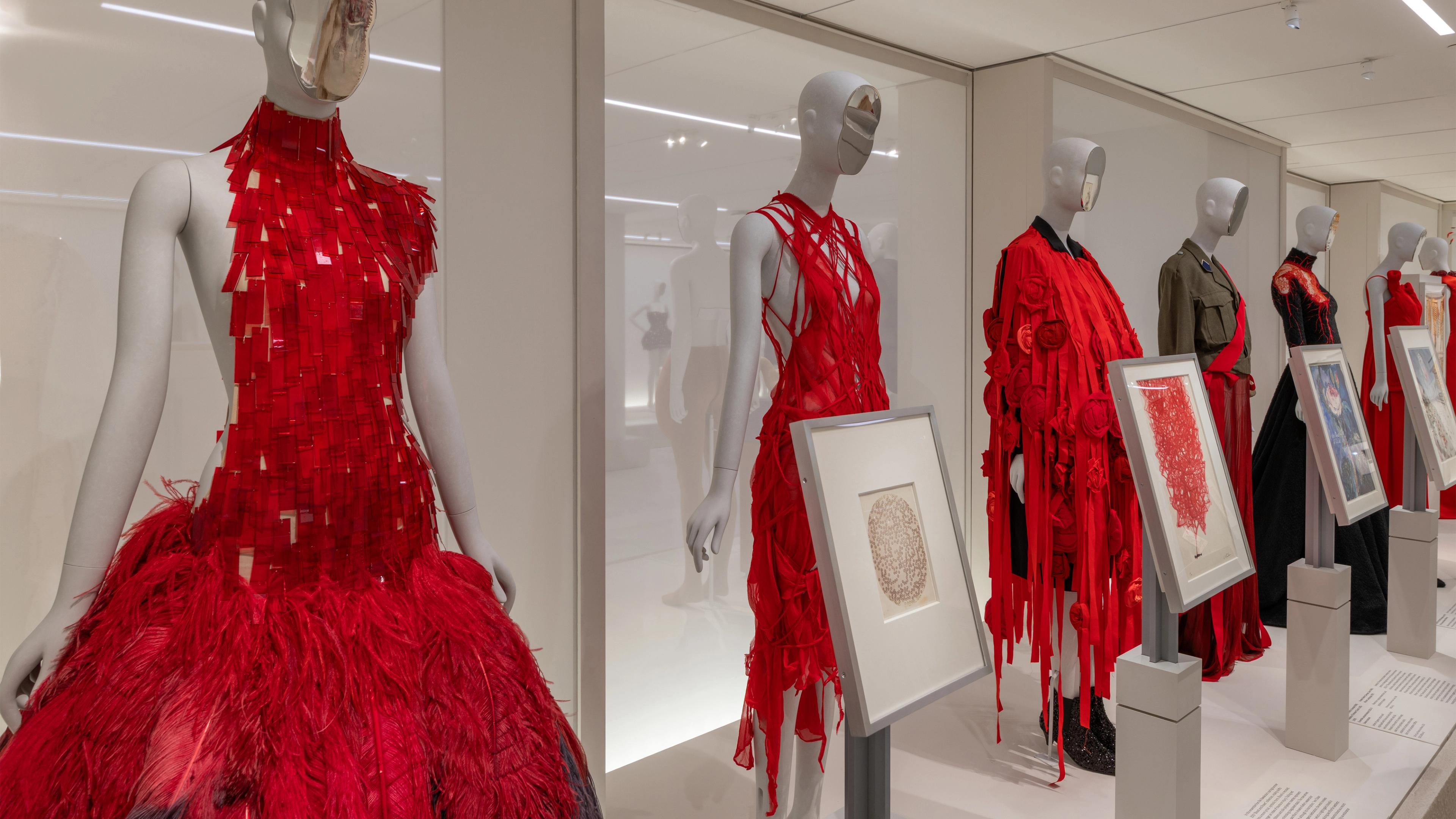 Mannequins in a fashion exhibit display vibrant red dresses with intricate textures of feathers and fabric strips, alongside framed art pieces on pedestals.