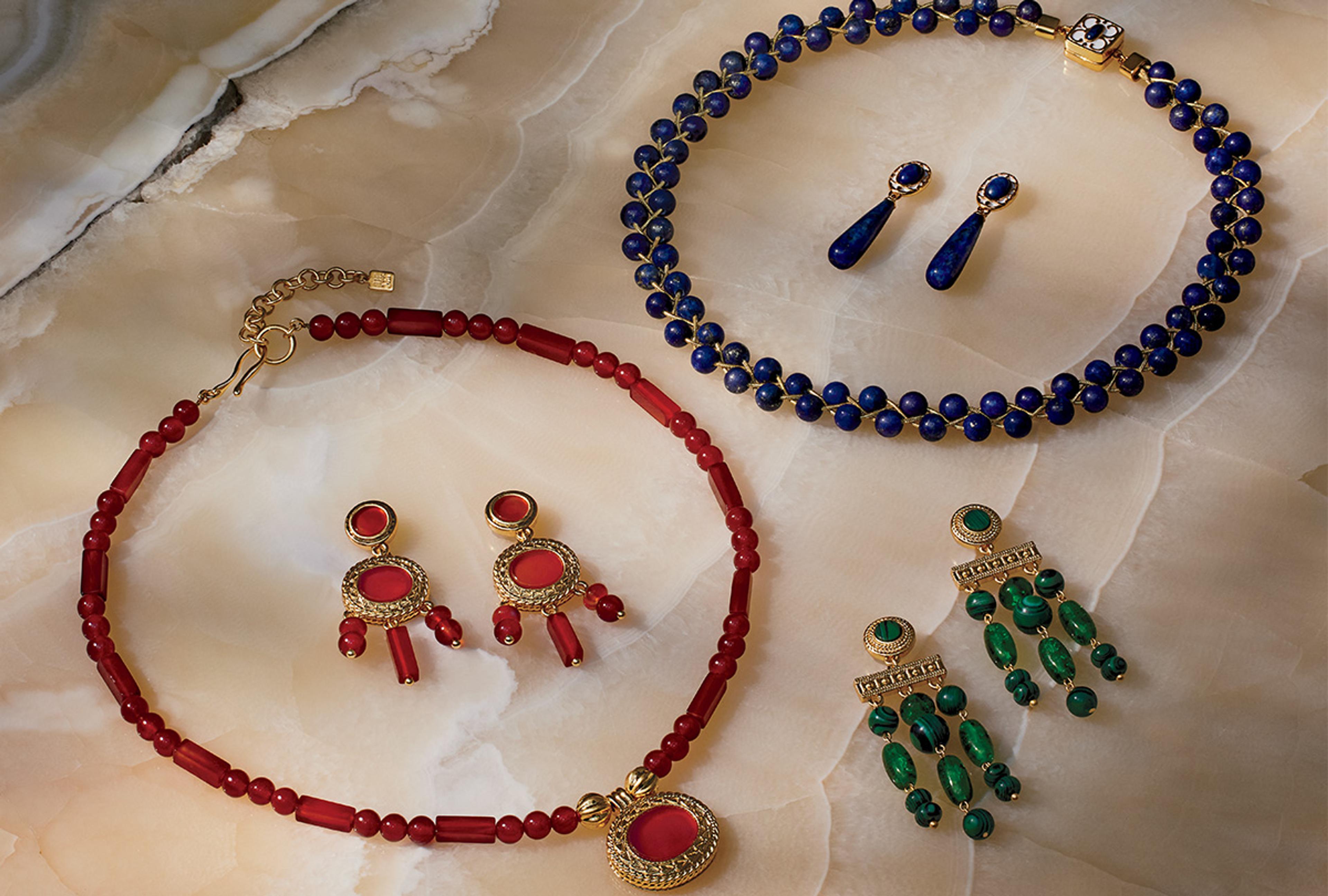 Three sets of earrings and two necklaces in red, blue, and green are displayed on a marble counter.
