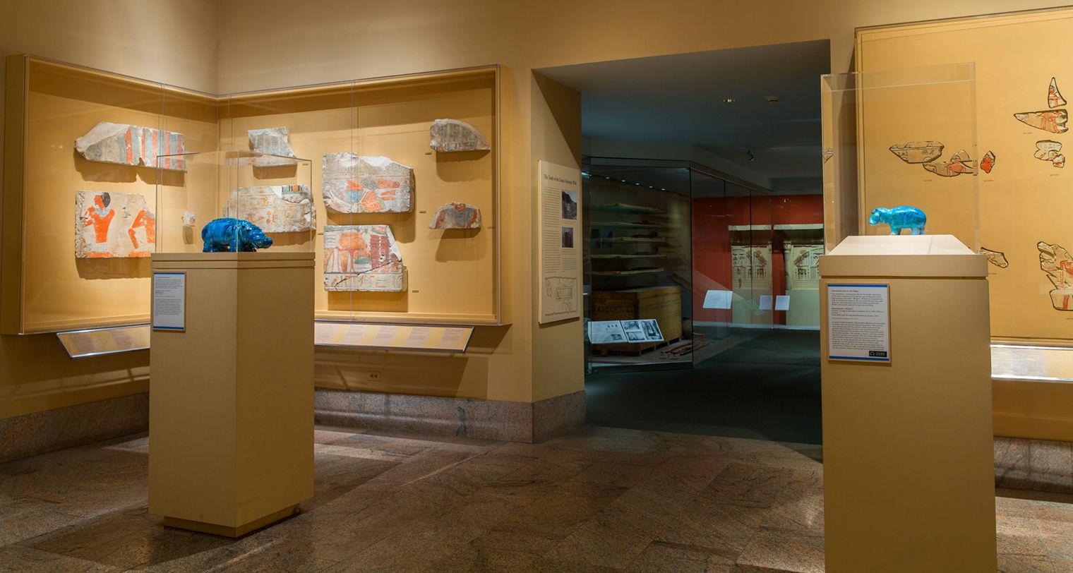 A museum gallery featuring two small blue hippos displayed on pedestals in glass cases with colorful stone reliefs hanging in the background.