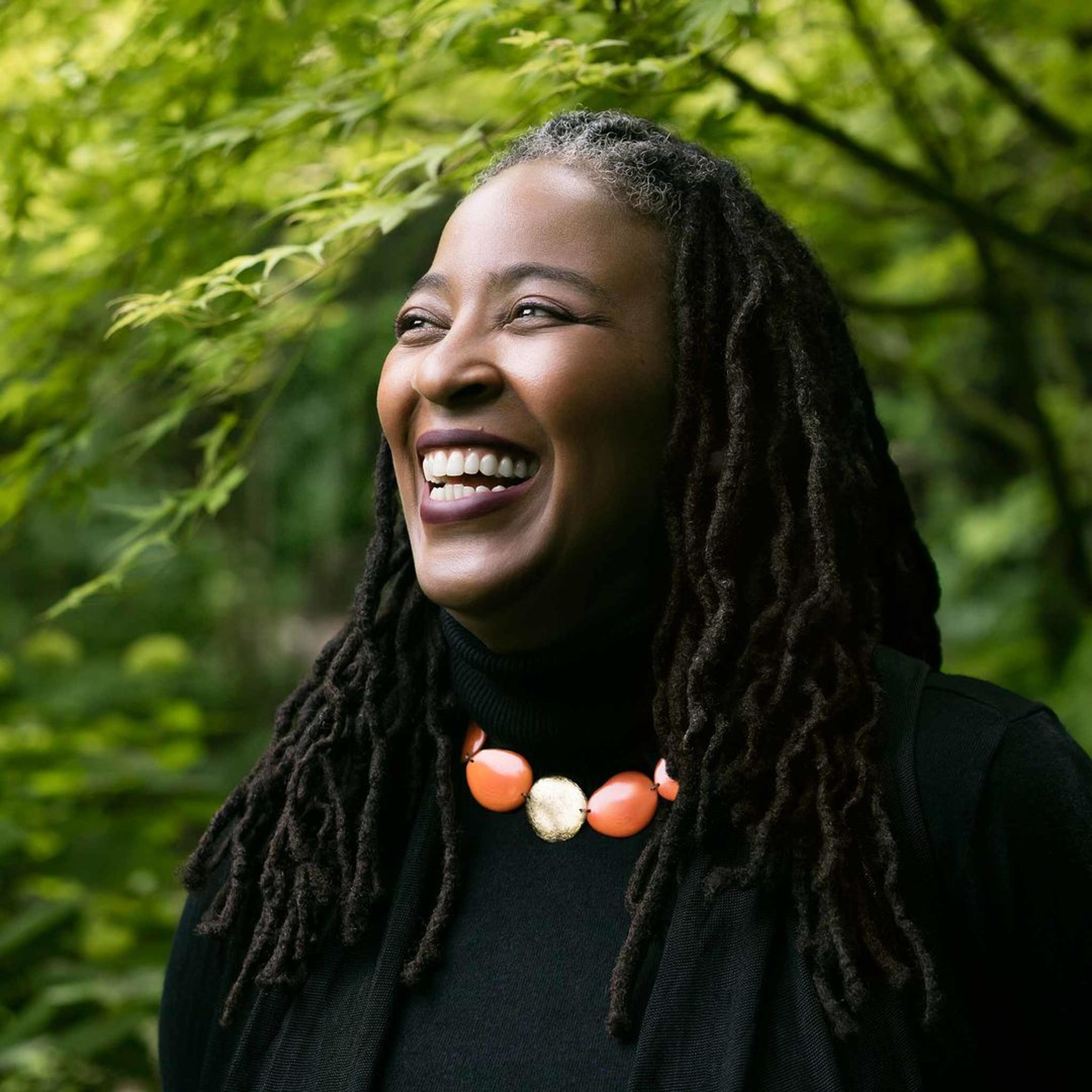 Poet Camille Dungy, a Black woman with long dreadlocks wearing a black turtleneck, laughing and standing among trees