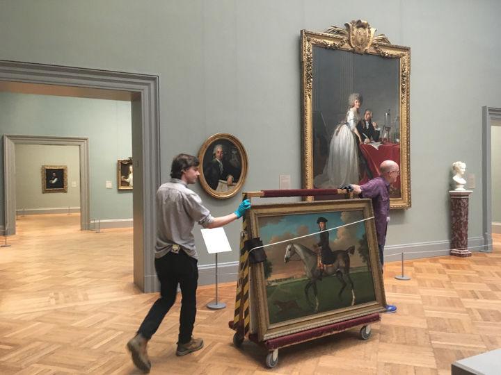 Gallery view showing a painting being moved by two Met technicians