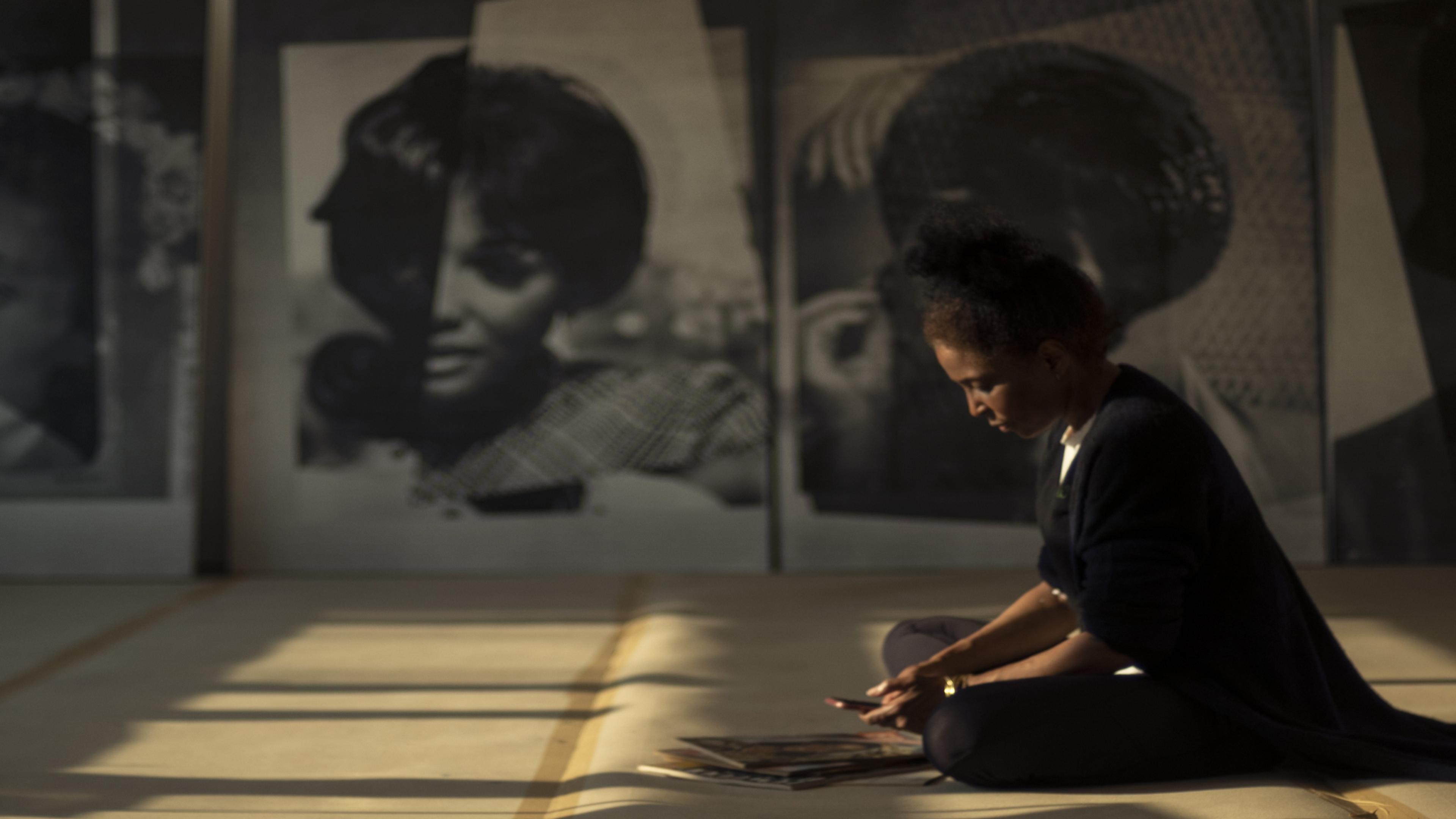 Lorna Simpson seated on the floor, bent over magazines. Rectangular panels of light illuminate the space, and large canvases with women's faces hang from the far wall.