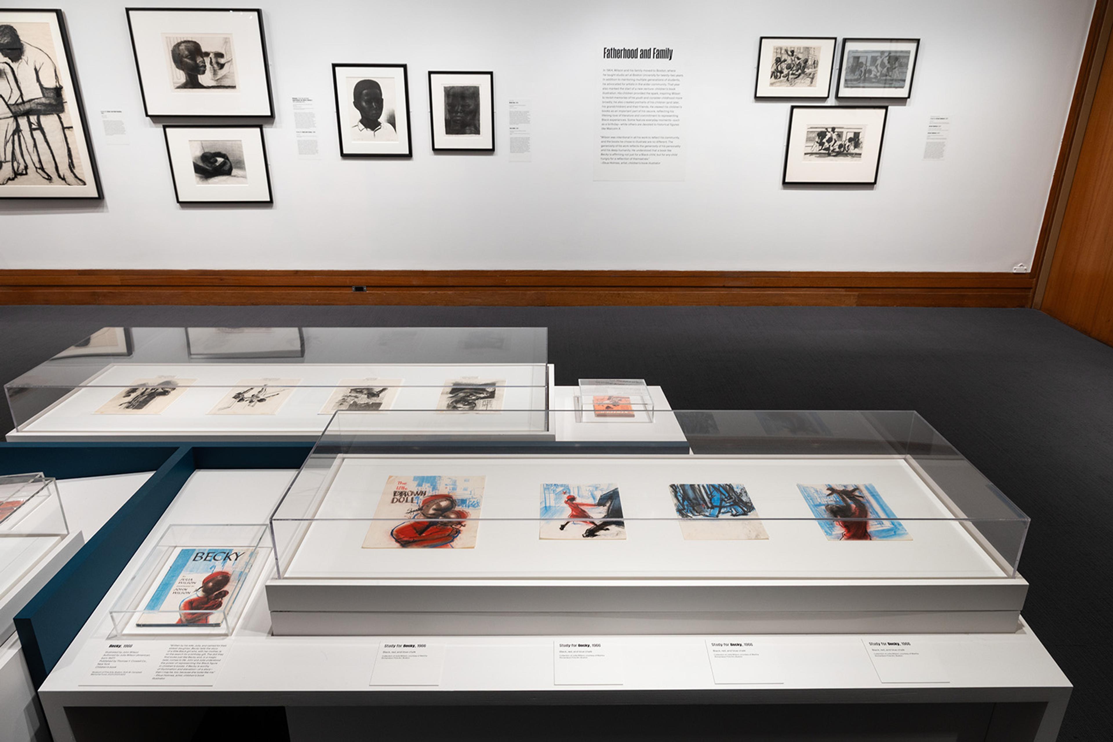 Art gallery exhibit featuring framed black and white and color artworks on walls and in glass cases.