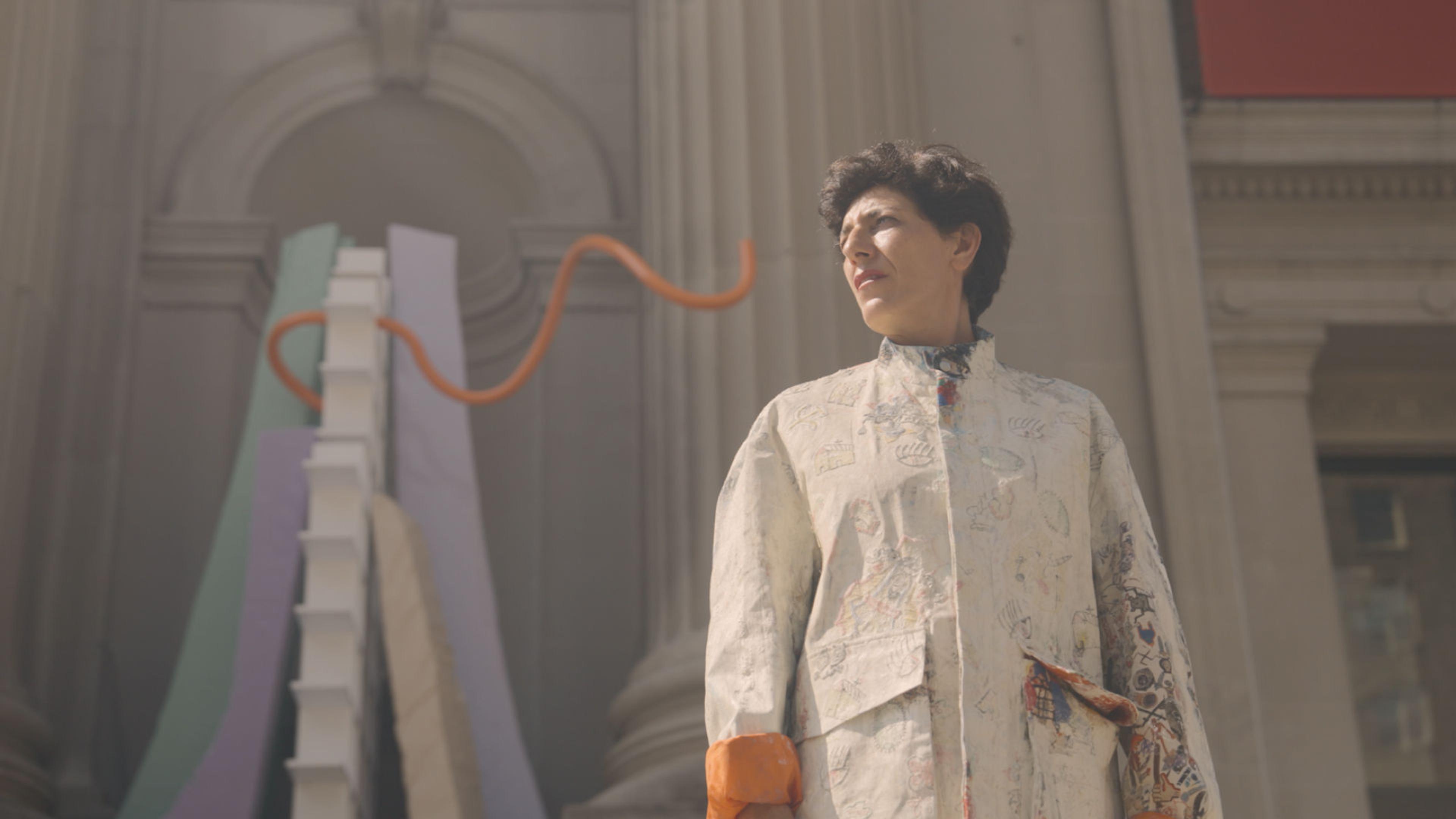 The artist Nairy Baghramian, with short brown hair and wearing a white jacket, stands before a large brightly colored modern sculpture installed in one of the niches on The Met's neoclassical facade.