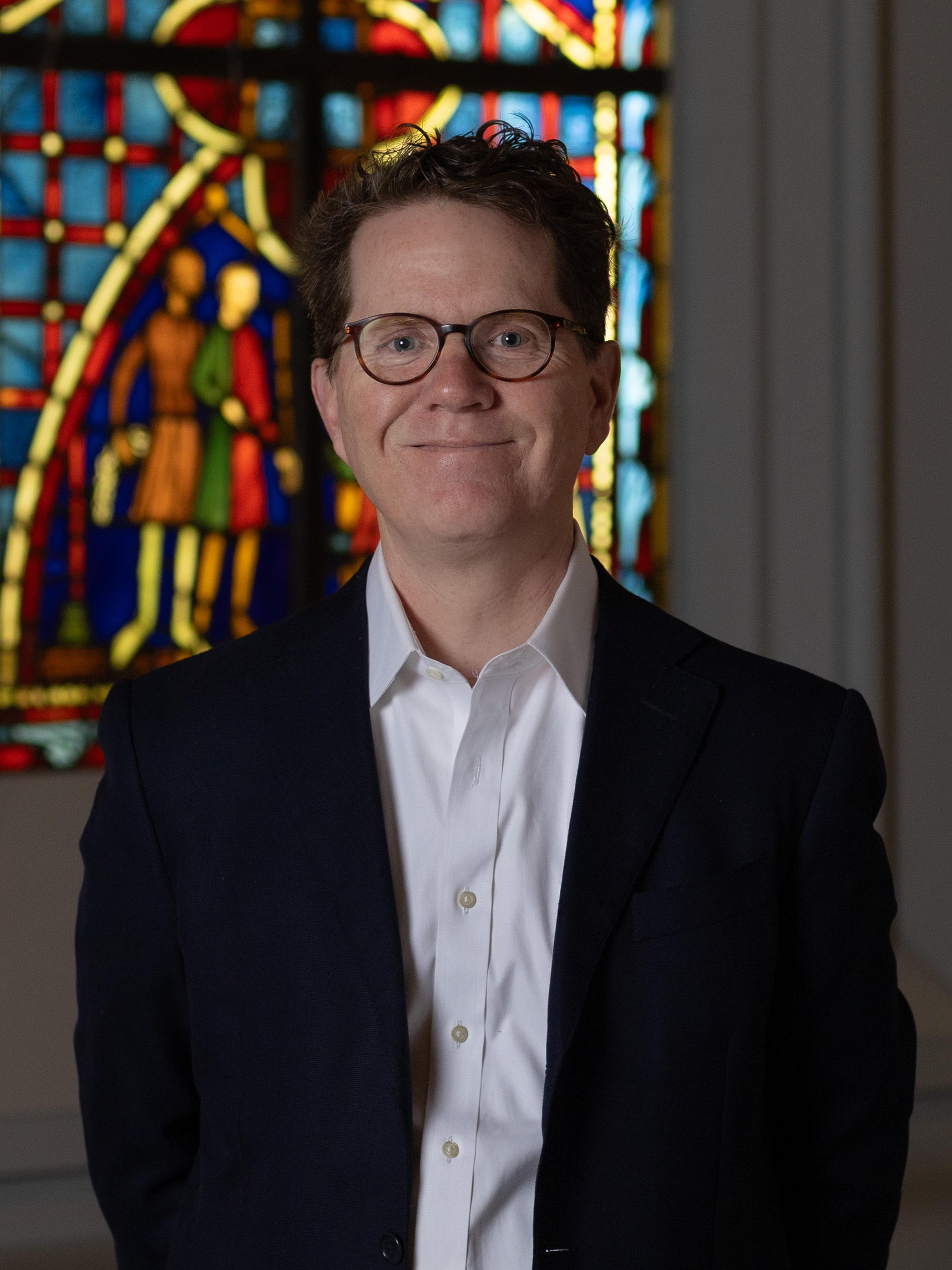 A man wearing a dark blazer and round glasses smiles in front of a stained glass window