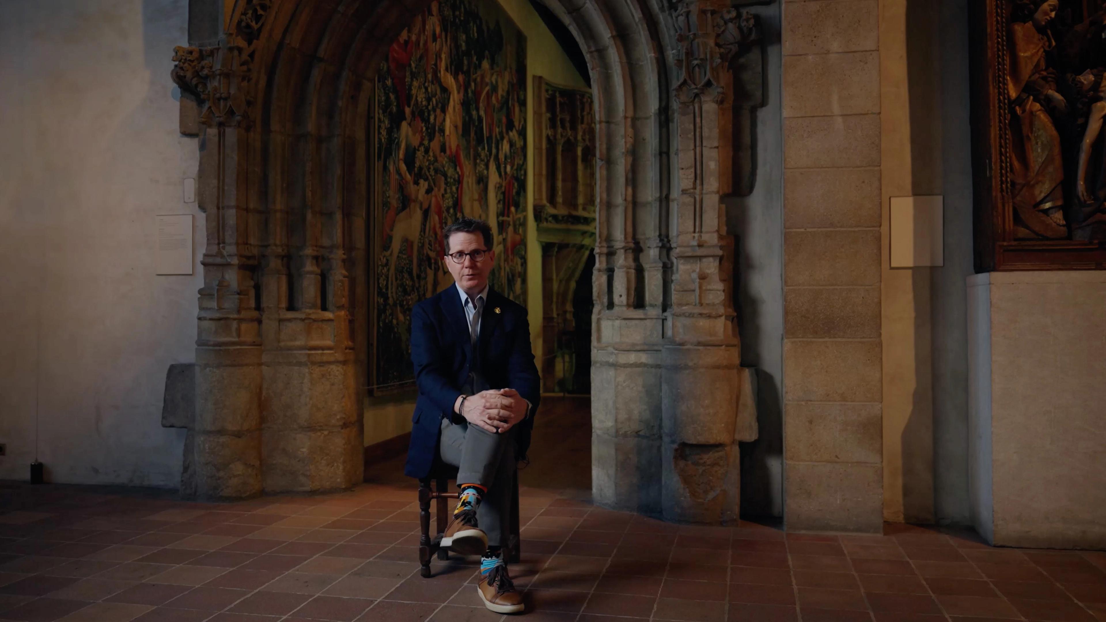 A bespectacled man in a navy suit sits in a wheelchair before an ornate Gothic arch and stained glass.