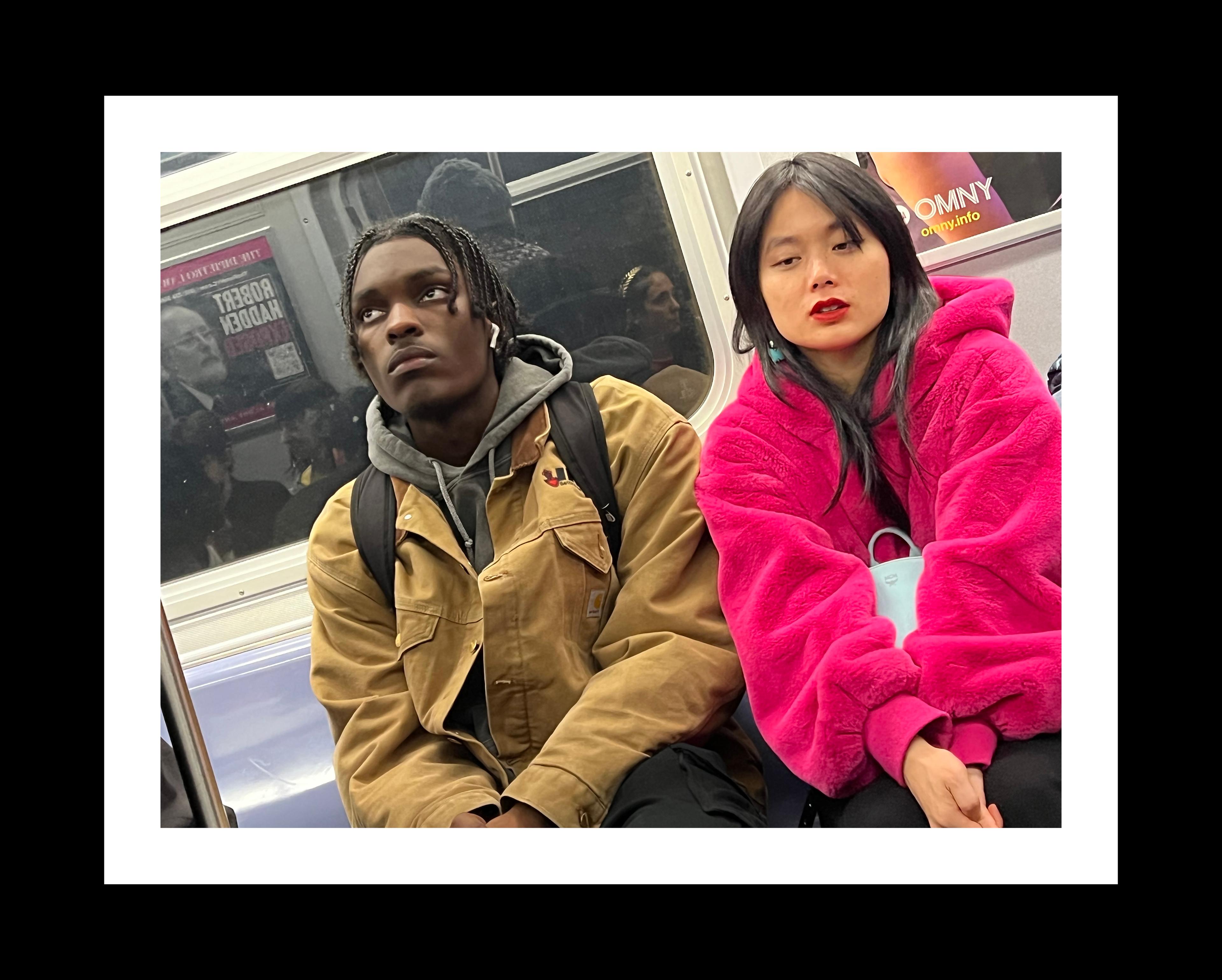 Color photograph of an adolescent Black male and Asian female sitting next to each other on a subway car. The male is looking up, the female is looking down. Both have their hands crossed in their laps. The Black teen has braided hair and wears a brown jacket with black pants and black shoulder straps from a knapsack behind him. The Asian teen has shoulder-length black hair and wears a fuzzy magenta coat with a white purse cradled between her arms.
