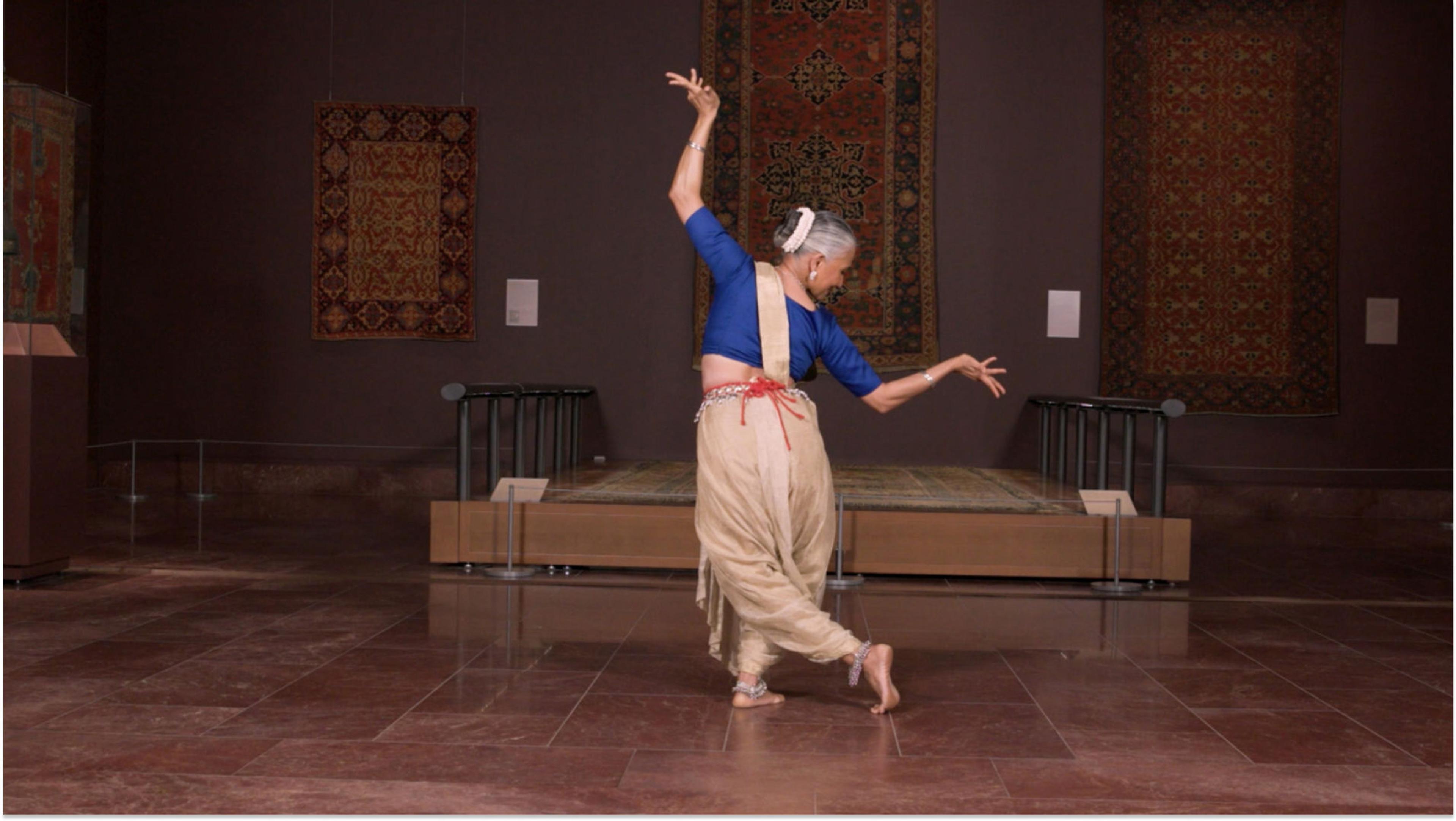 A woman wears a blue top, tan bottoms, a tan strap diagonally around her torso, and a red rope around her waist. She stands with her shoulders tilted down towards the right. Her arms are bent at a 90 degree angle and her hands are flexed. Her left leg is bent and crossed behind her right. Behind her are rugs hanging on the wall.