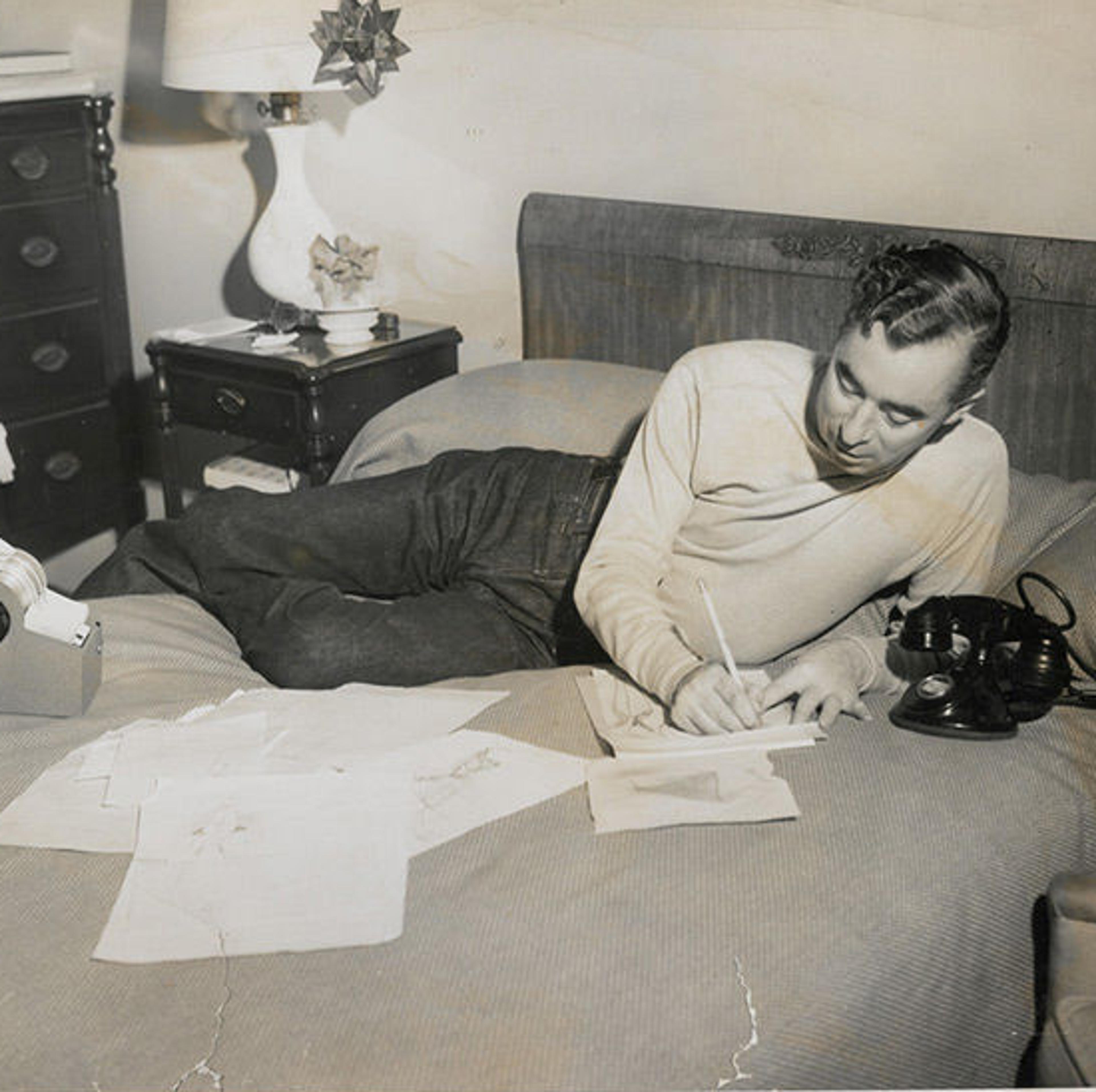 Charles James reclines on a bed strewn with papers. He takes notes on a notepad.