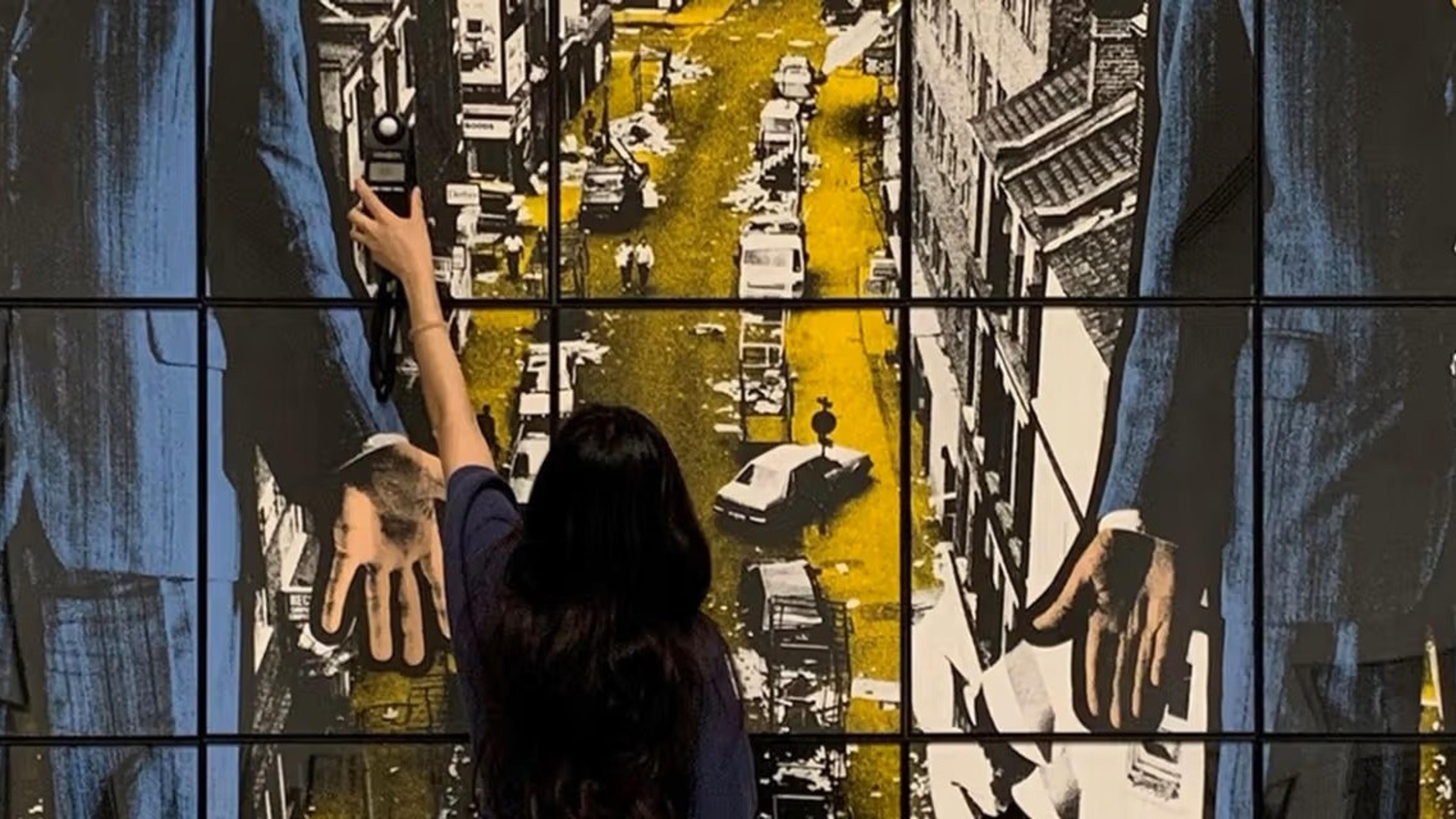 A woman stands facing an artwork with an image of a yellow street in the center and two people standing in blue suits on the side.