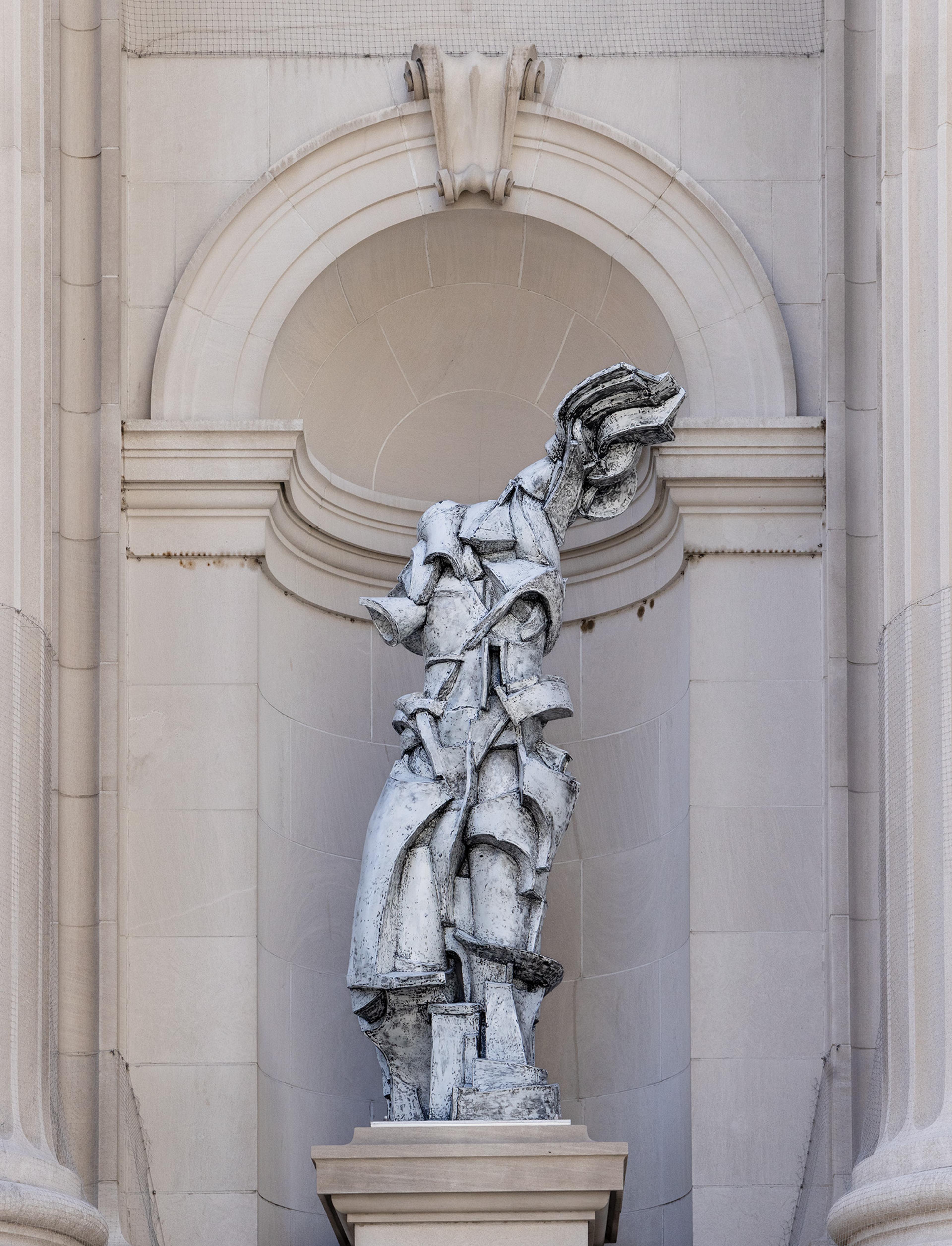 A modern, abstract stone statue of a draped figure stands in an arched alcove on a classical building facad