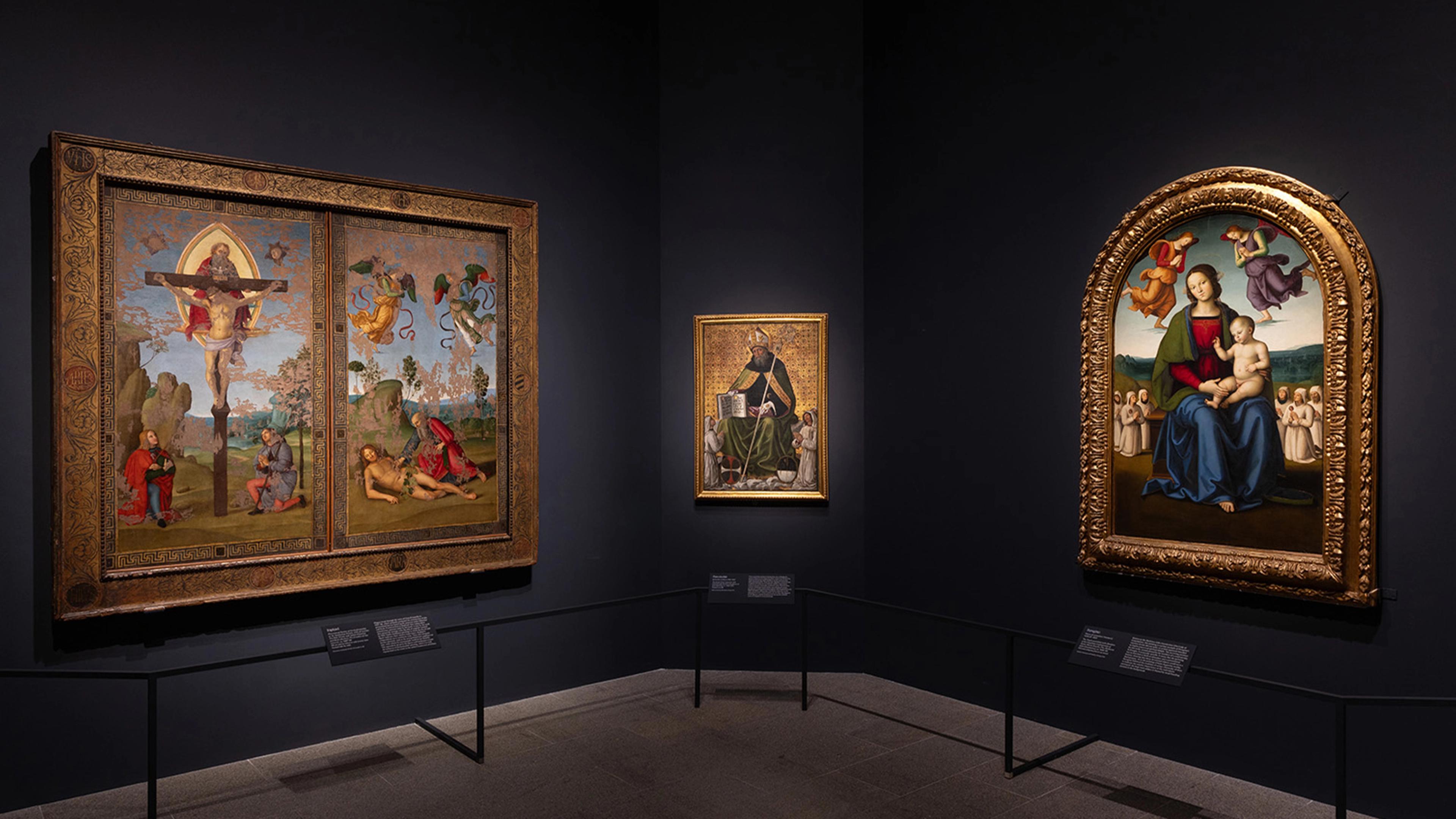 A dimly lit art gallery features three framed Renaissance paintings on dark walls.
