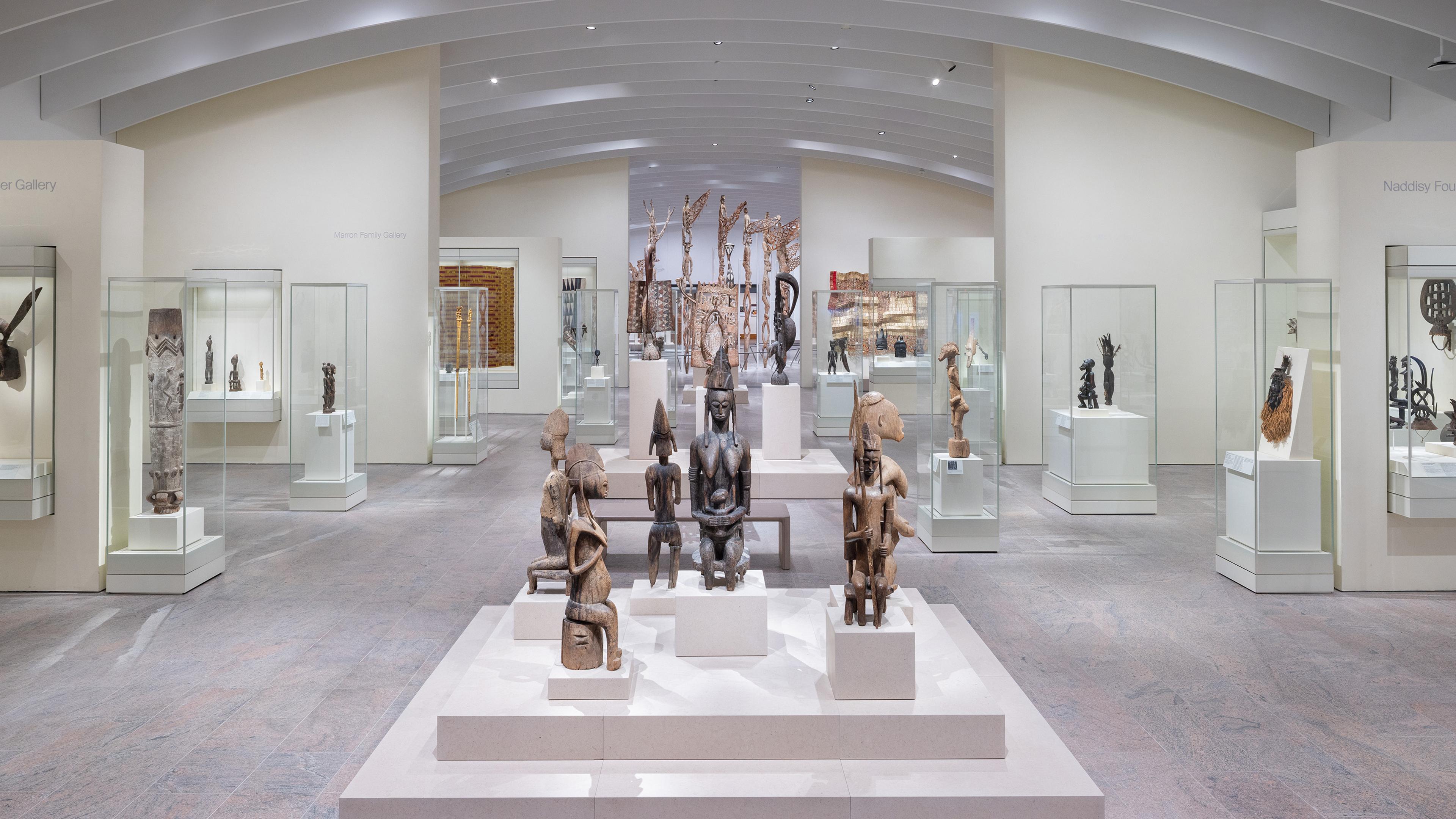 A gallery of wooden sculptures in glass cases under a vaulted ceiling