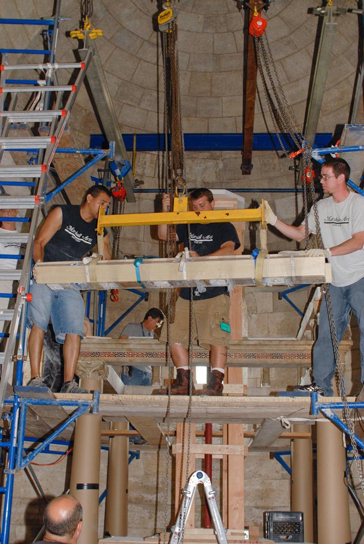 Riggers lowering a lintel down from the scaffolding. The original stone lintel is inside a protective covering of wooden planks.